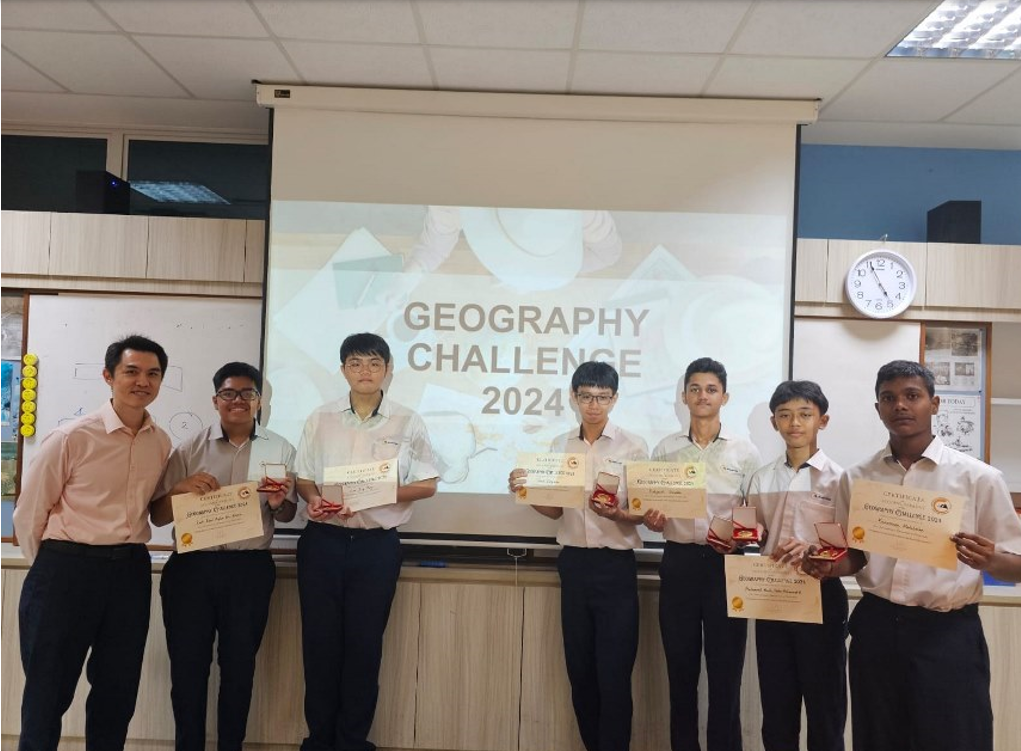 Seven people stand in a classroom holding certificates and medals in front of a Geography Challenge 2024 projection screen.