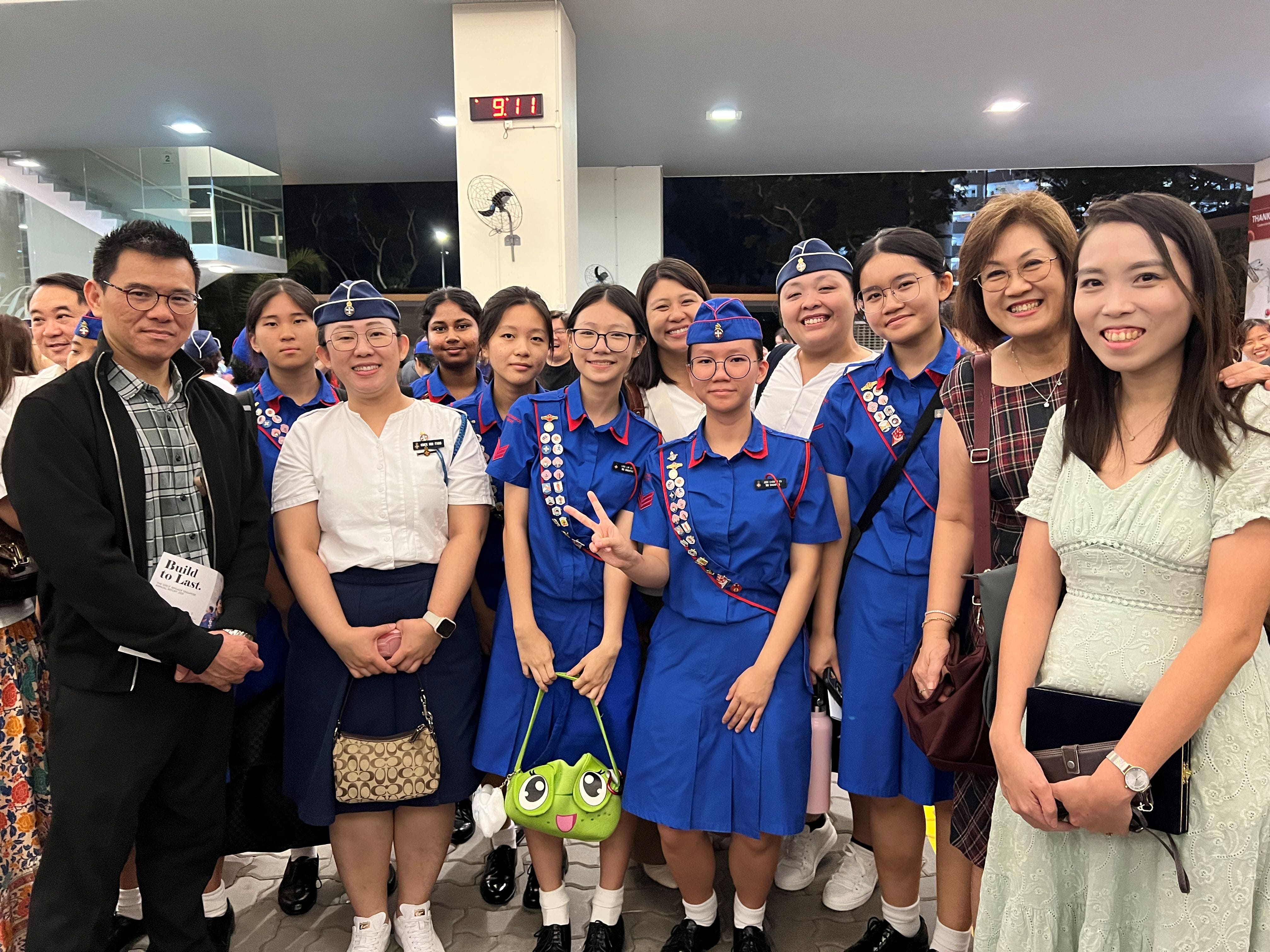 A group of smiling young women in blue uniforms with badges pose with adults indoors at 9:11 PM.
