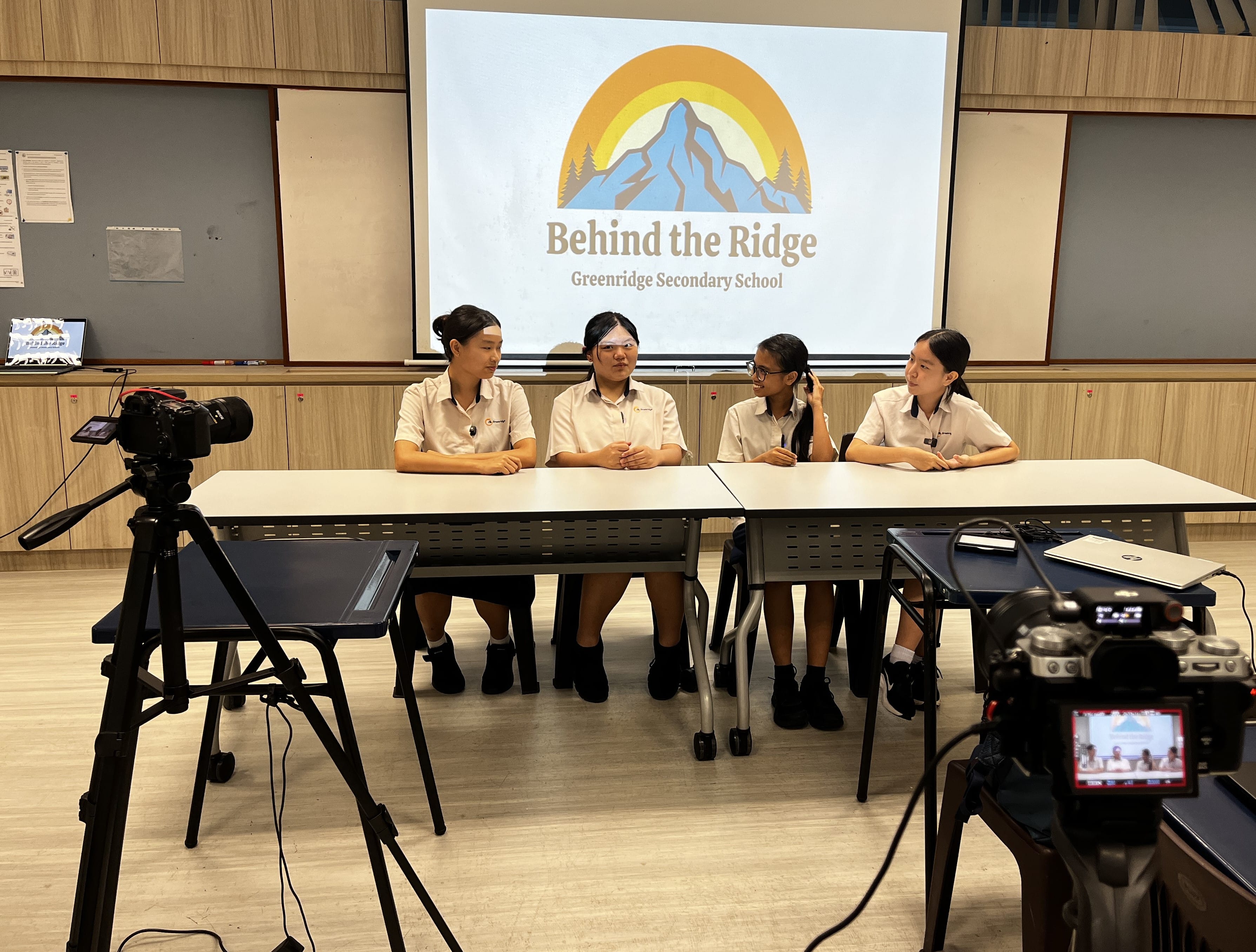 Four female students in uniforms sit at a table being filmed, with a Behind the Ridge Greenridge Secondary School screen behind them.