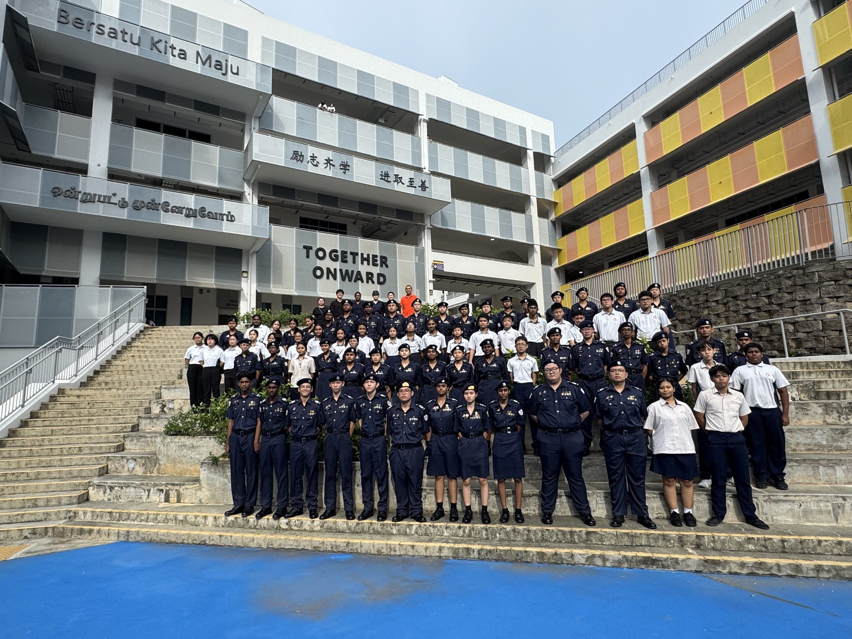 Large group of students in navy uniforms posing on outdoor steps in front of a school building reading Together Onward.