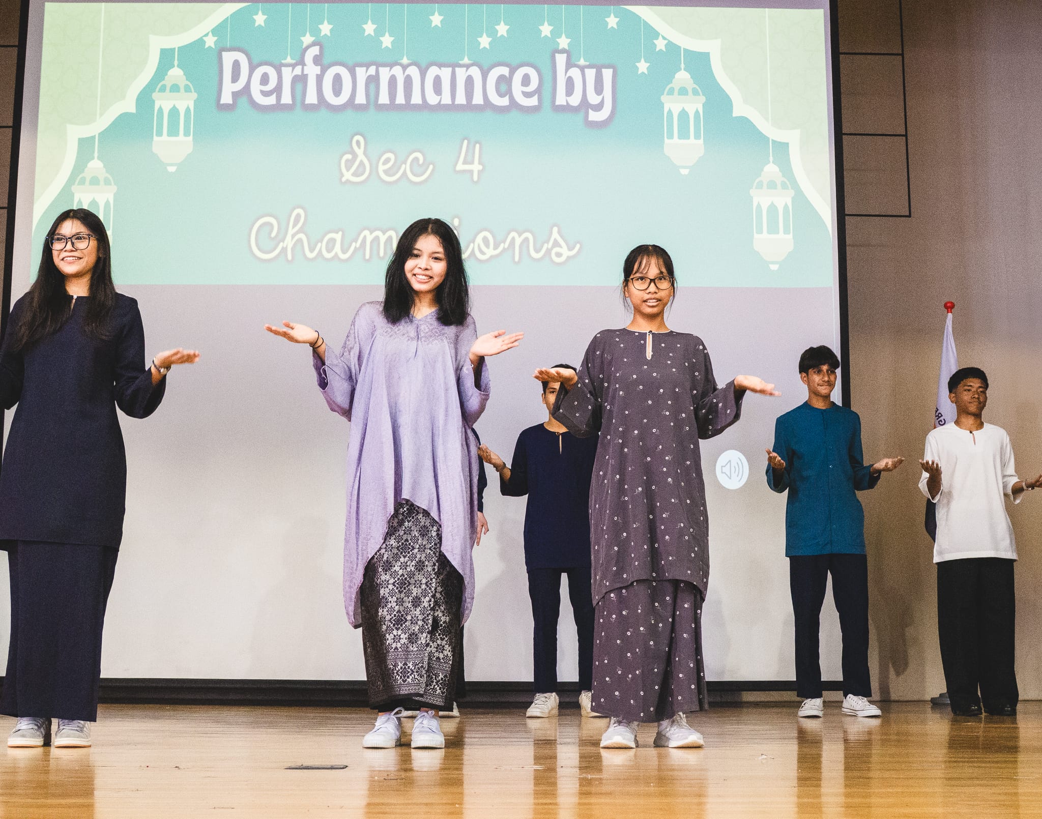 Six students perform on stage in traditional attire before a screen reading "Performance by Sec 4 Champions."