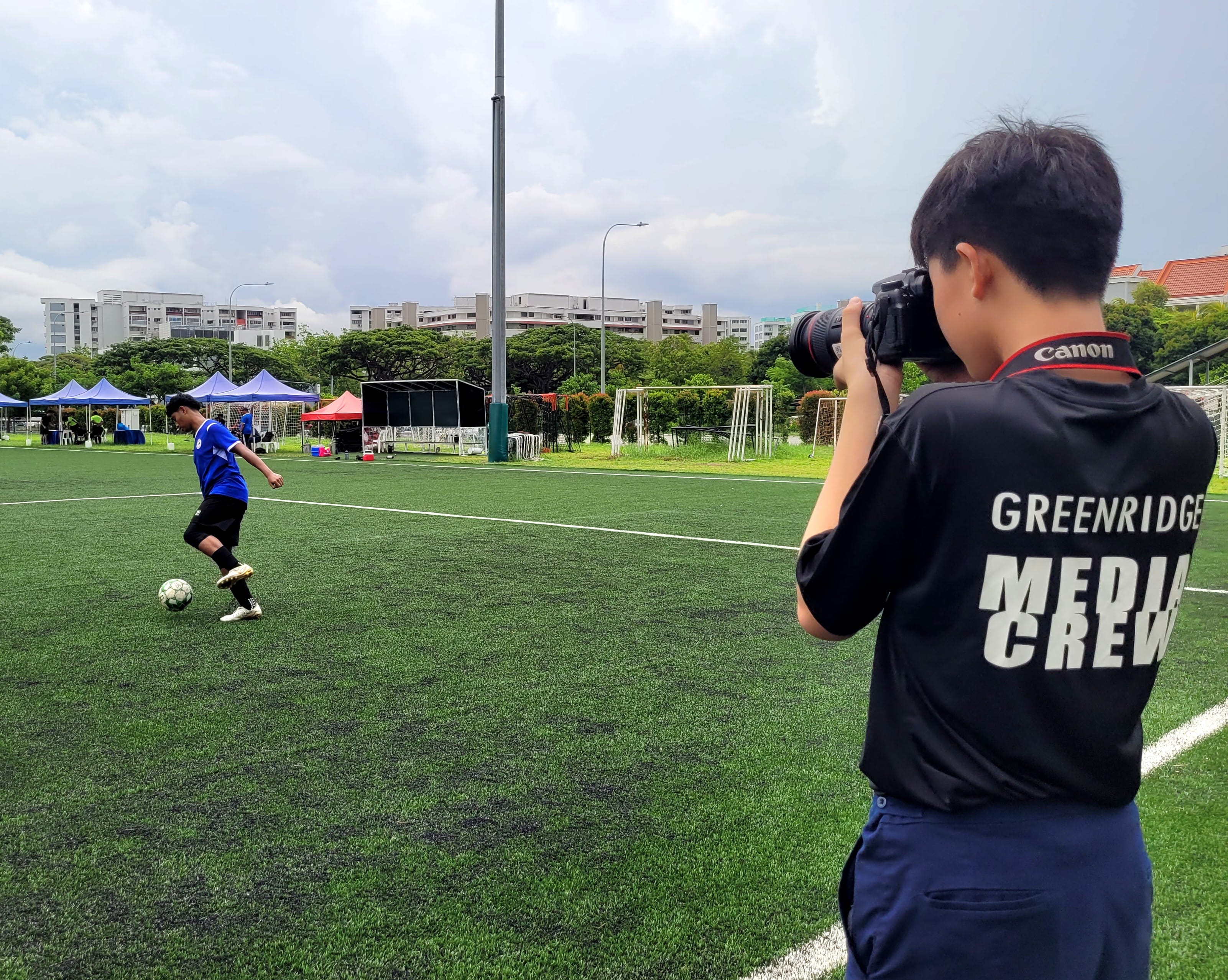 A Greenridge Media Crew member photographs a soccer player dribbling a ball on a green turf field.