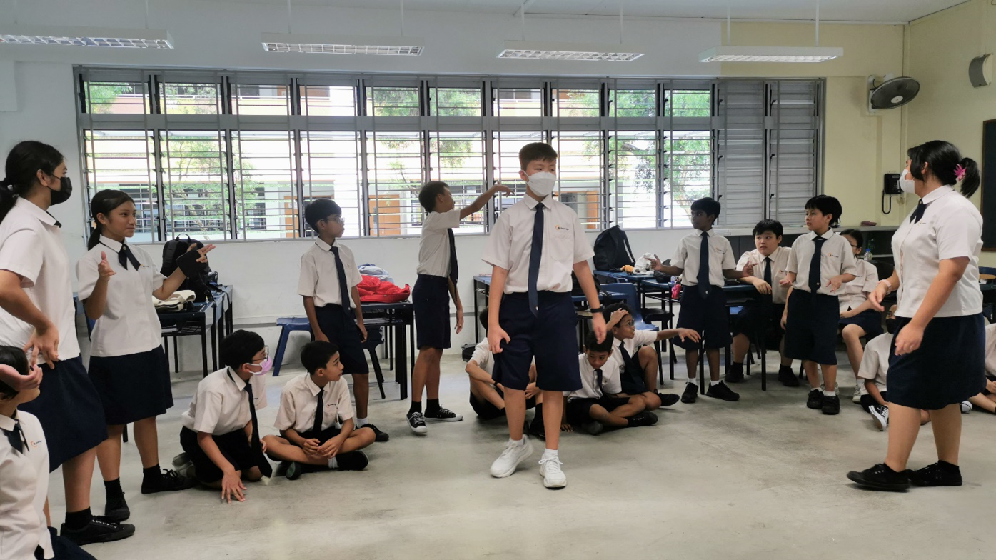 Students in white shirts and navy uniforms stand and sit in a classroom during an active group activity.