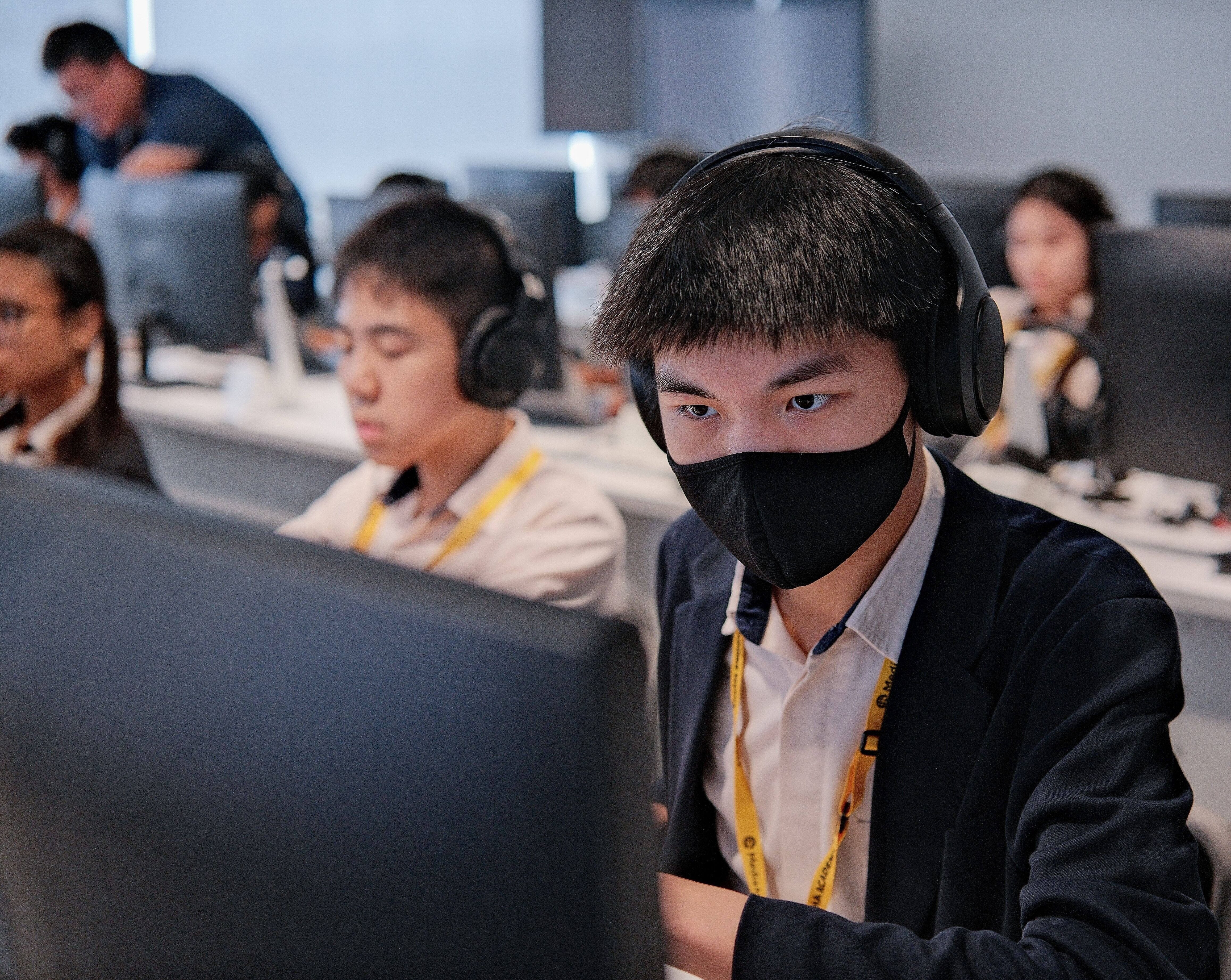 Student in mask and headphones focuses on a computer in a classroom with other students at monitors behind.