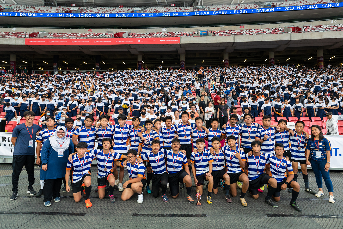 Youth rugby team wearing blue and white striped jerseys with medals pose at 2023 National School Games stadium.