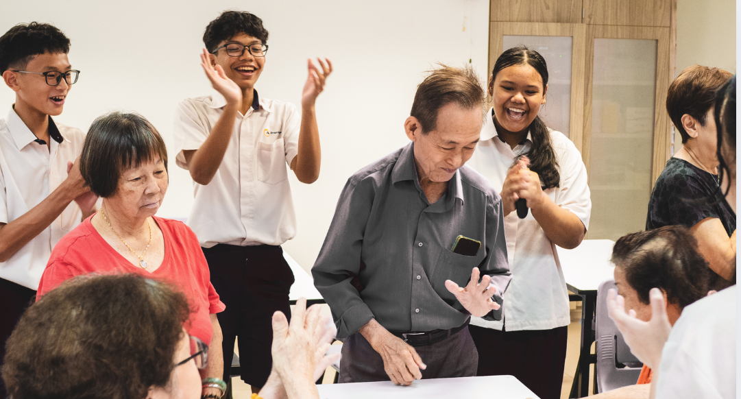 A group of young people clapping and cheering around an elderly man standing at a table indoors.
