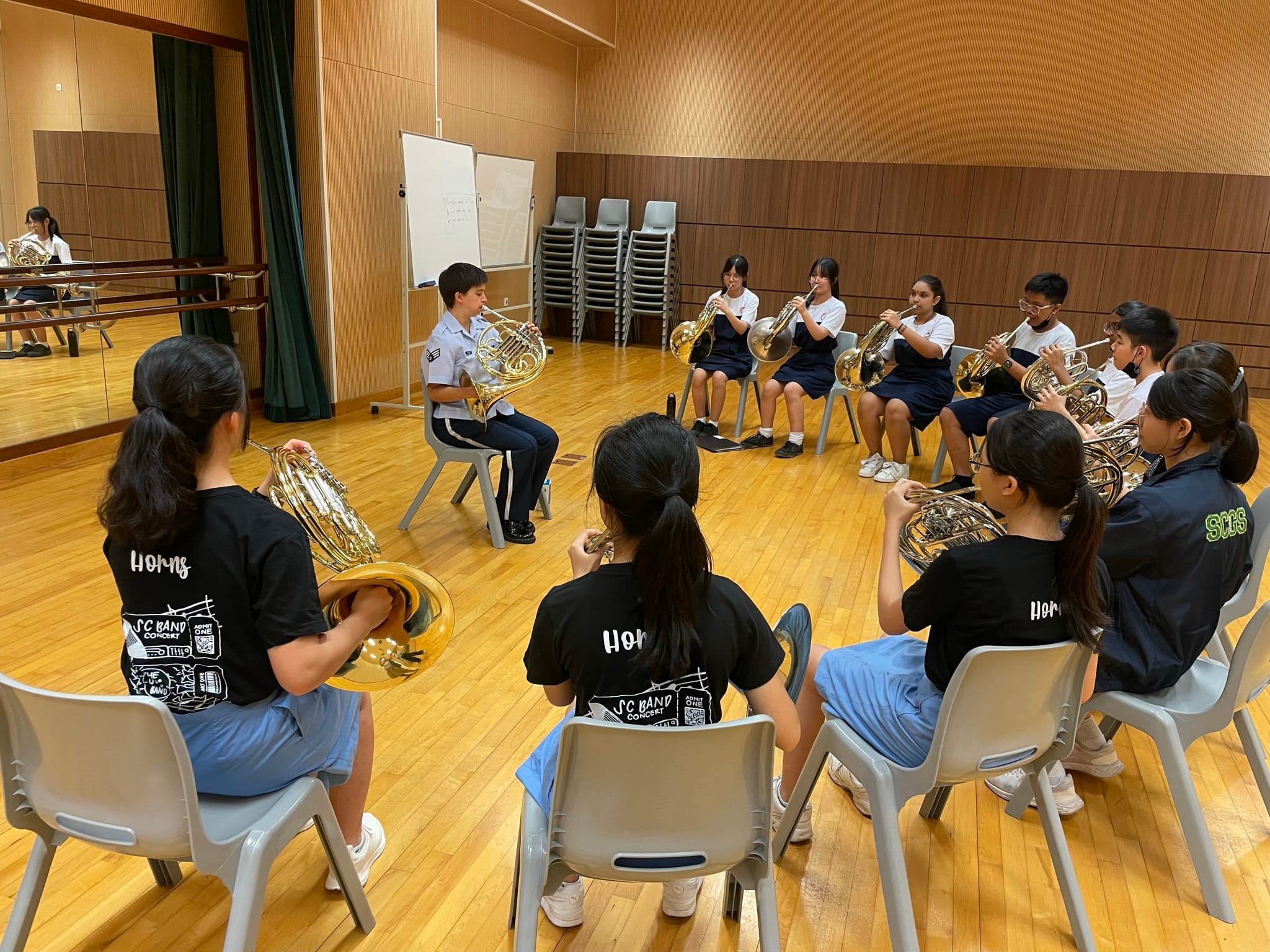 Students sit in a circle on chairs in a rehearsal room, playing French horns led by an instructor.