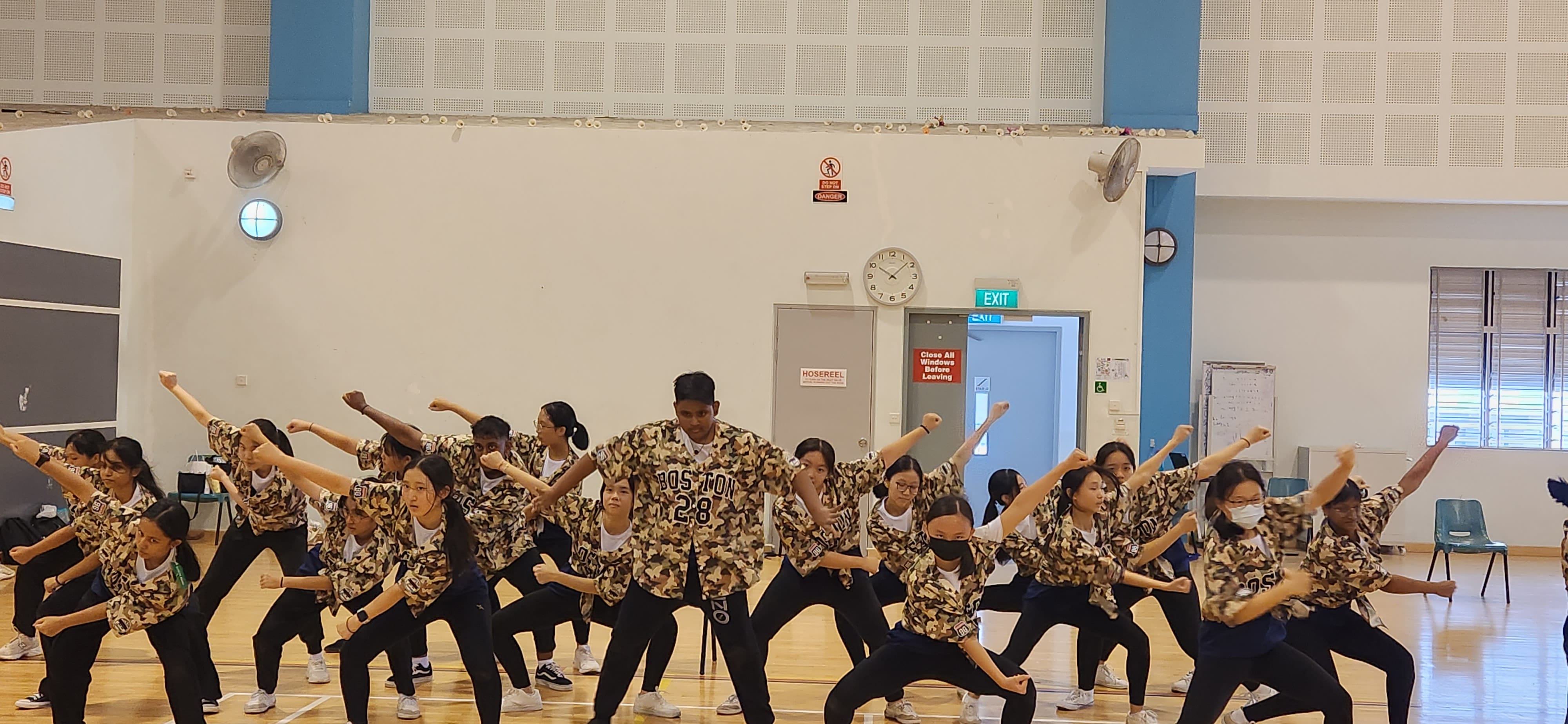 A large group of students in camouflage jerseys perform a synchronized dance routine in a school gymnasium.