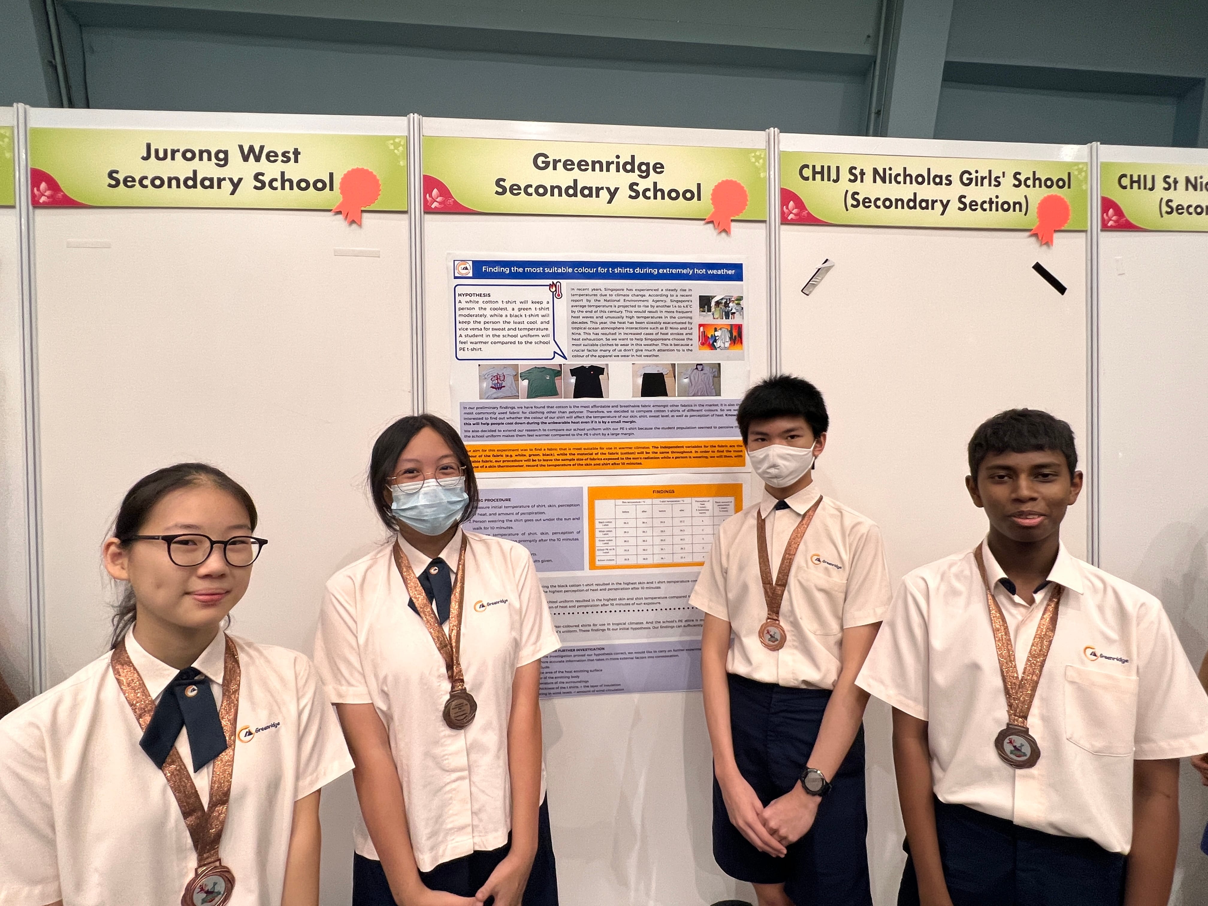Four students wearing bronze medals stand before a Greenridge Secondary School science fair poster about t-shirt colors in hot weather.