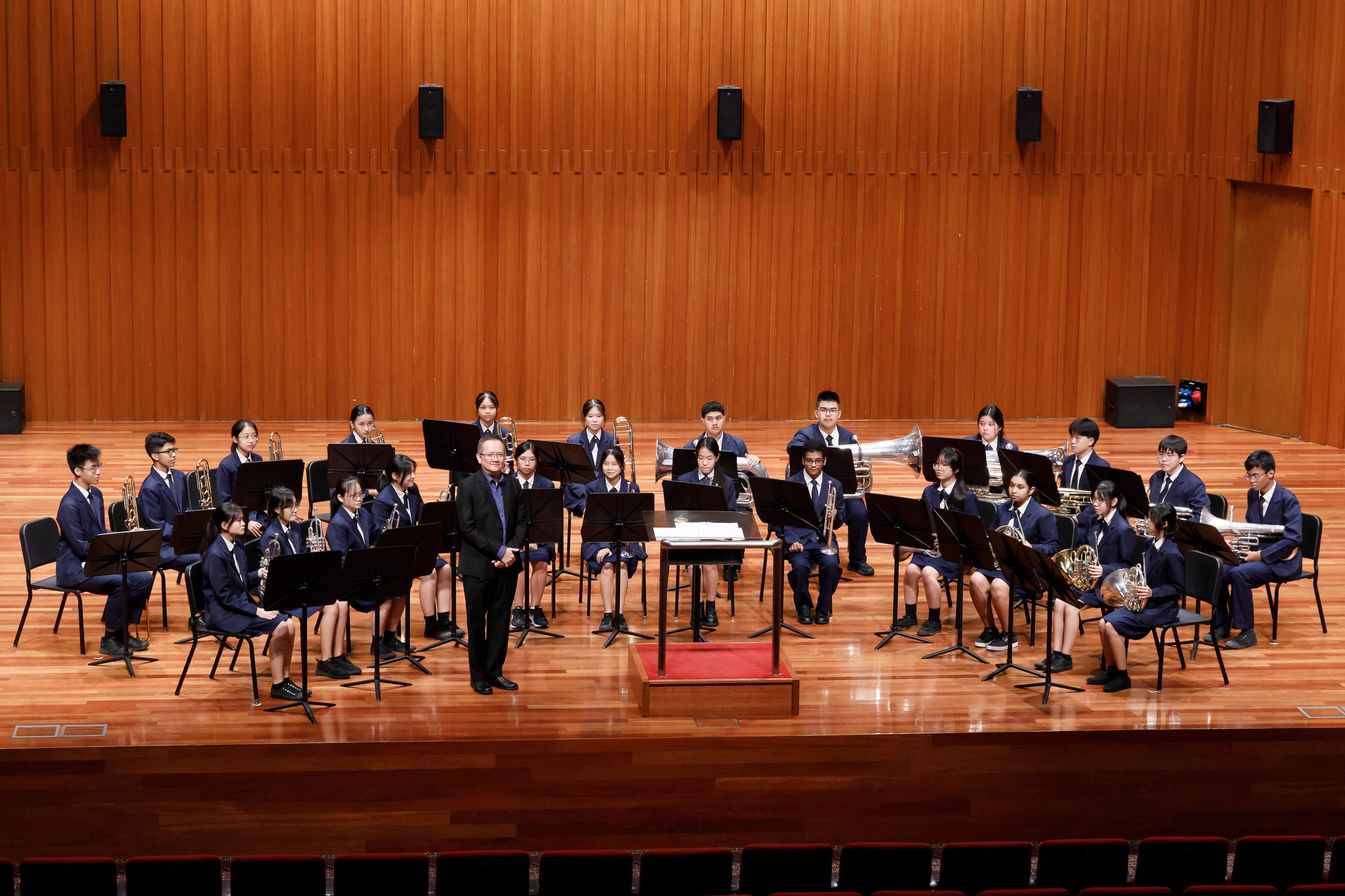 Students in navy uniforms sit with brass instruments on a wood-paneled stage, with a conductor standing at center.