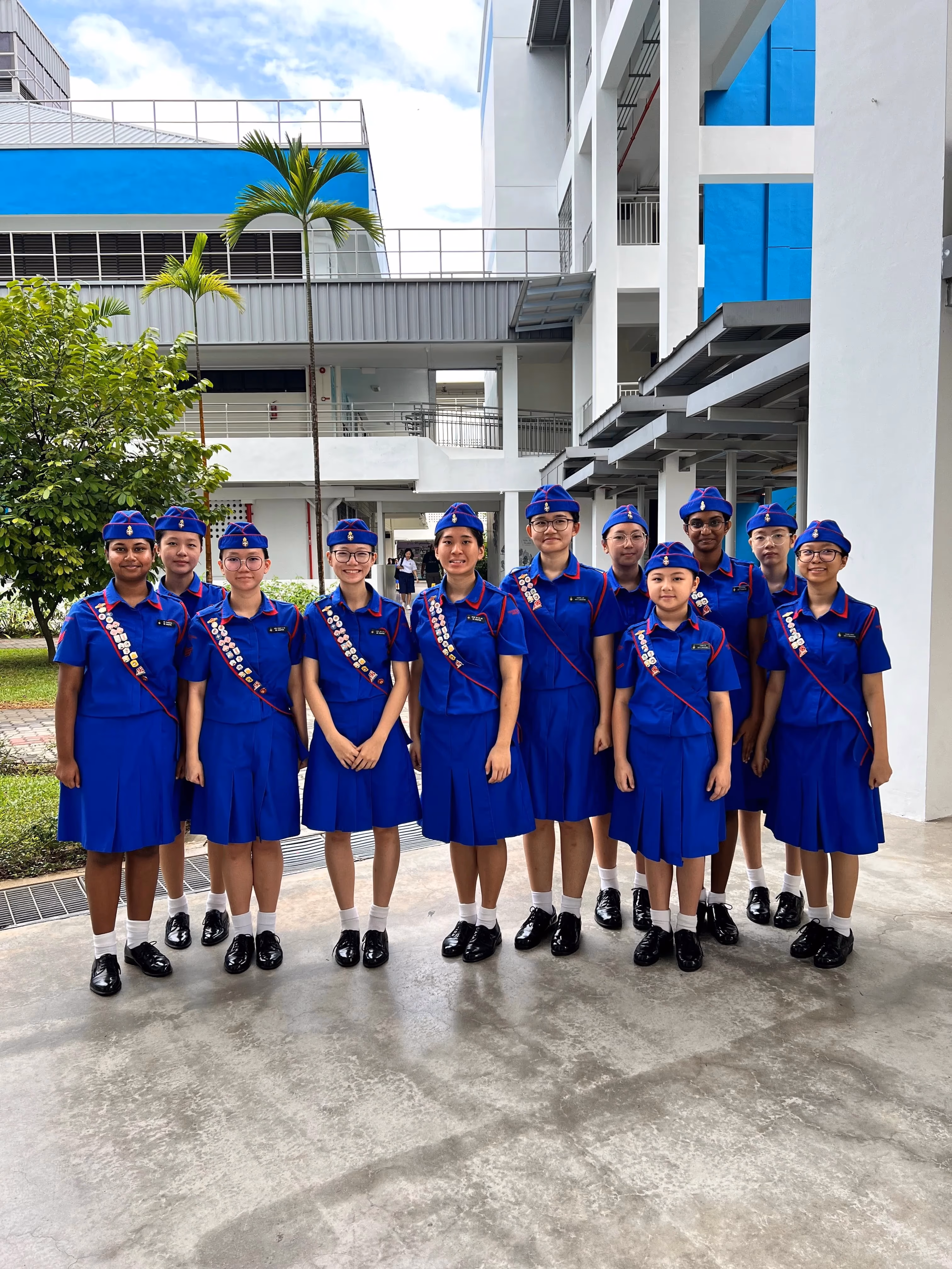 Eleven girls in blue uniforms with caps and badge sashes stand together outdoors near a school building.