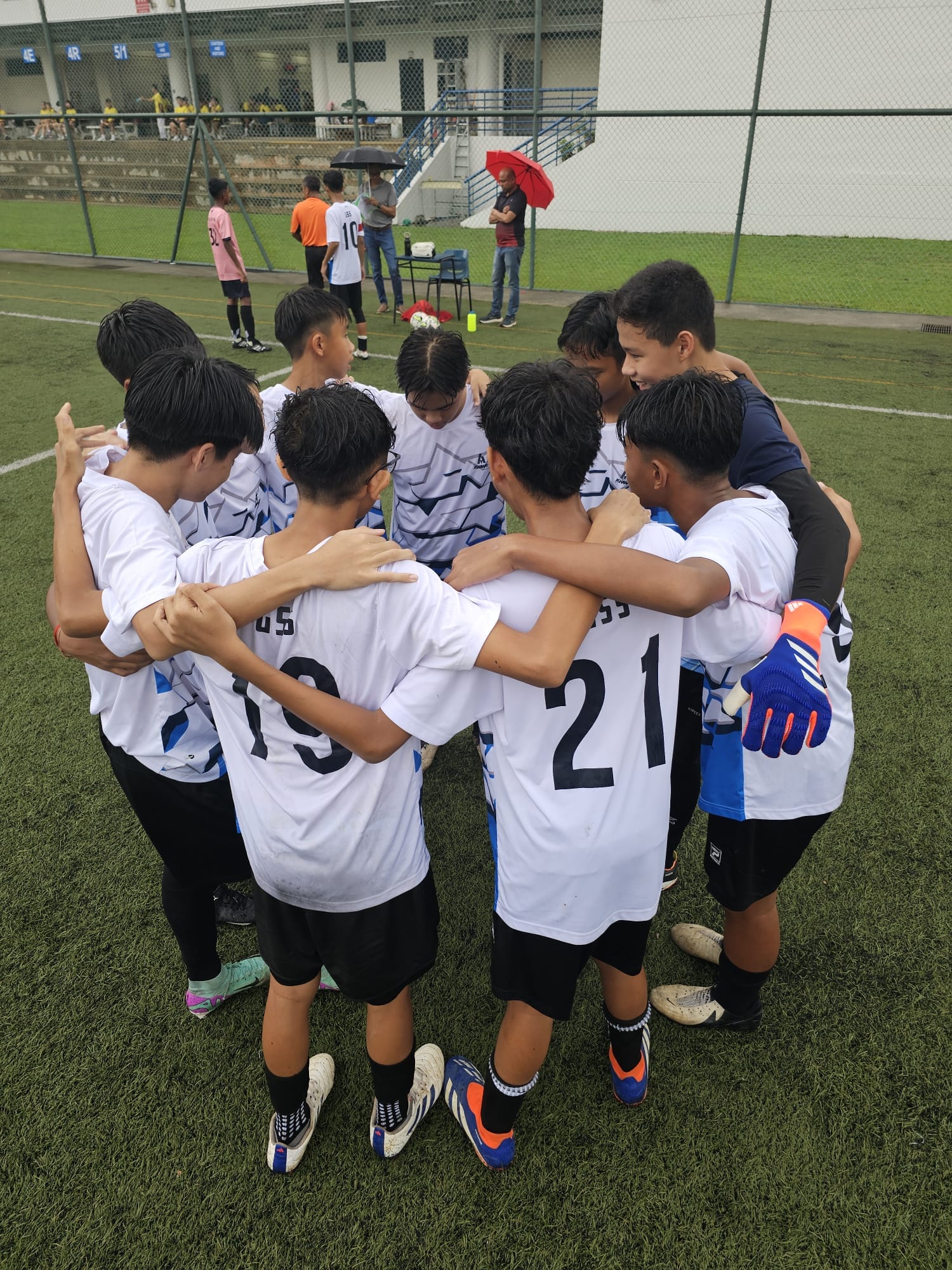 Young football players in white numbered jerseys huddle together on a green pitch near a stadium.