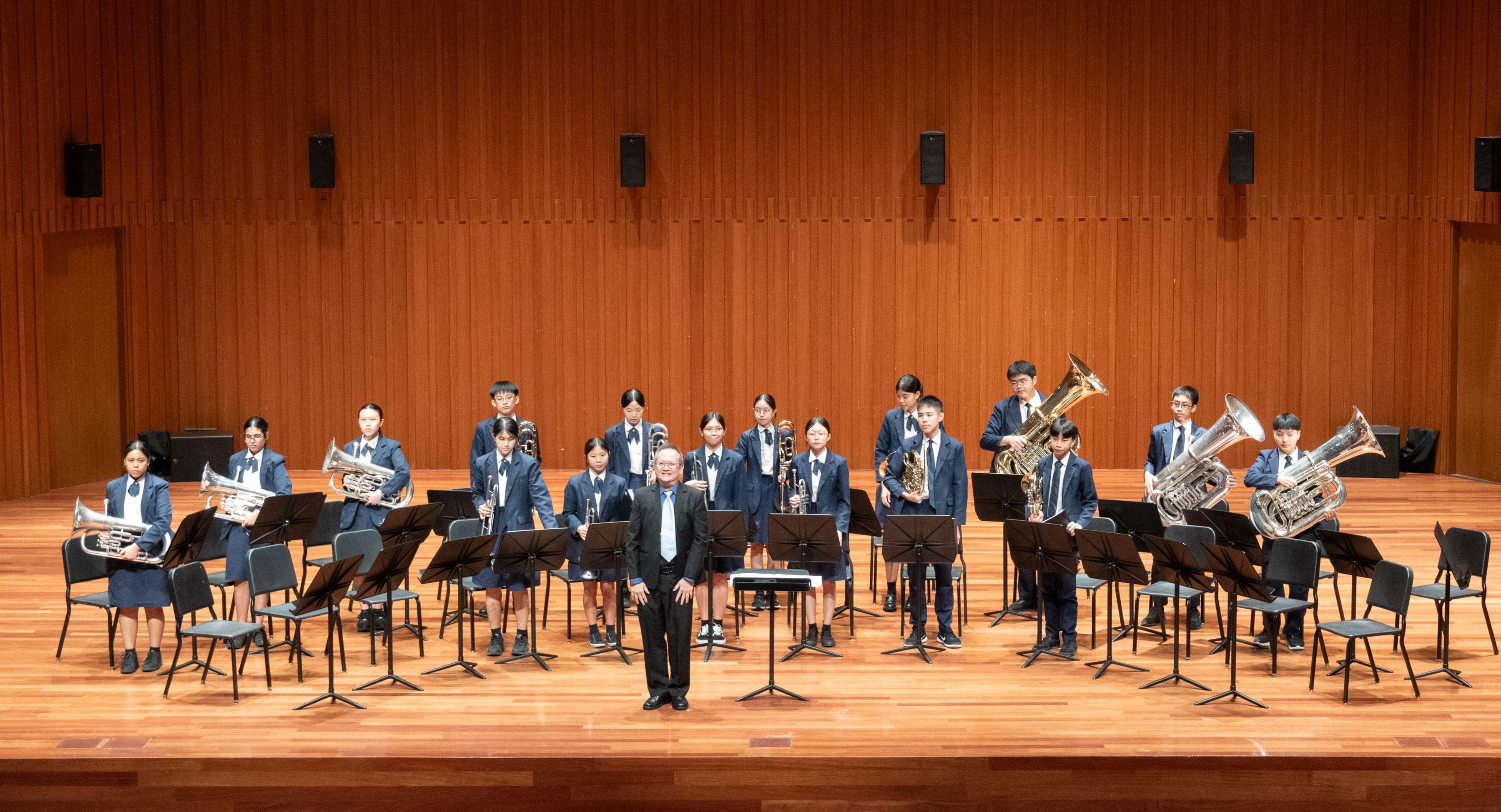 A youth brass band in blue uniforms poses on a wooden stage with an adult conductor standing at the center.