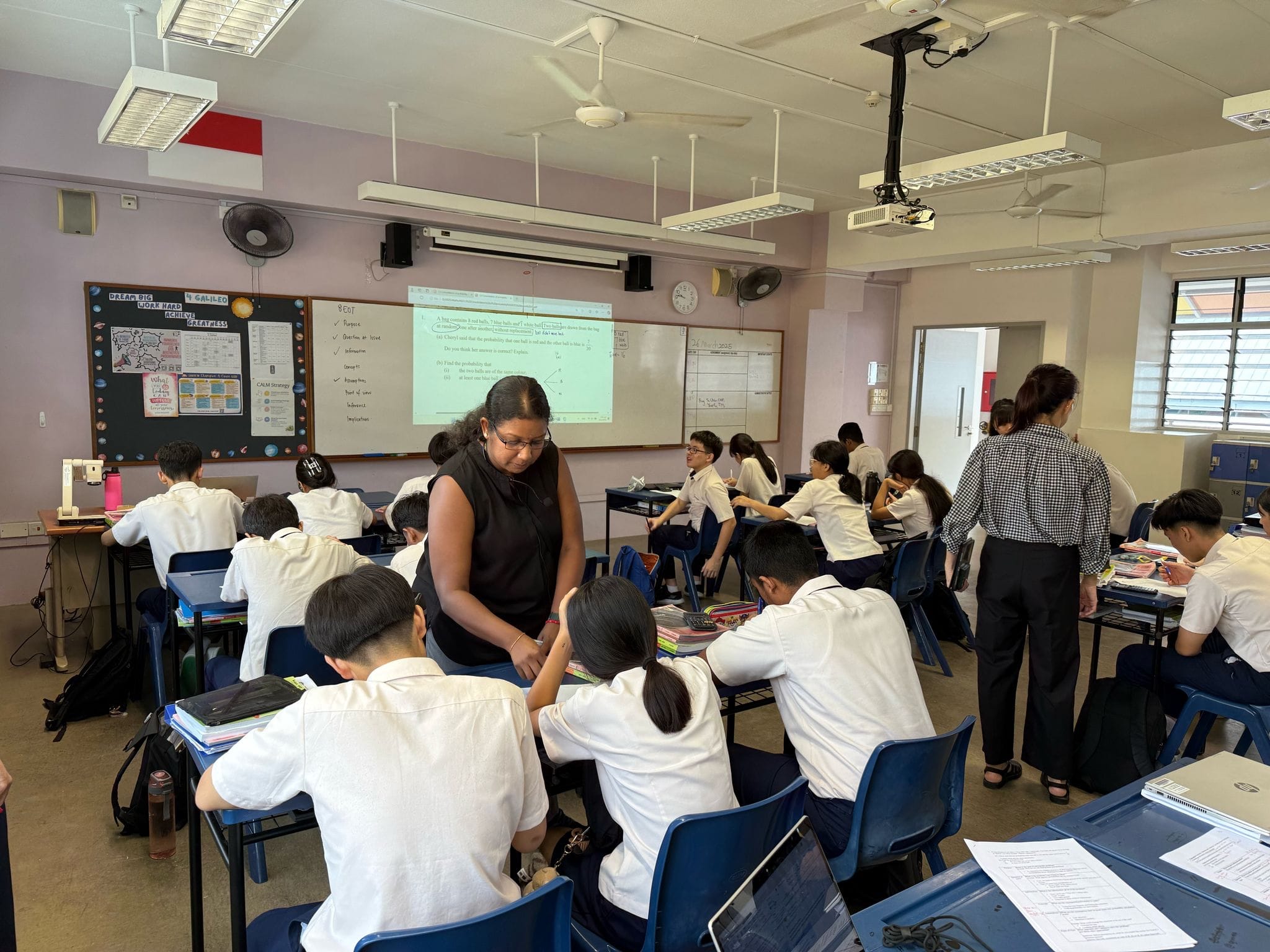 Two teachers assist uniformed students seated at blue chairs in a classroom with a projected lesson on a whiteboard.