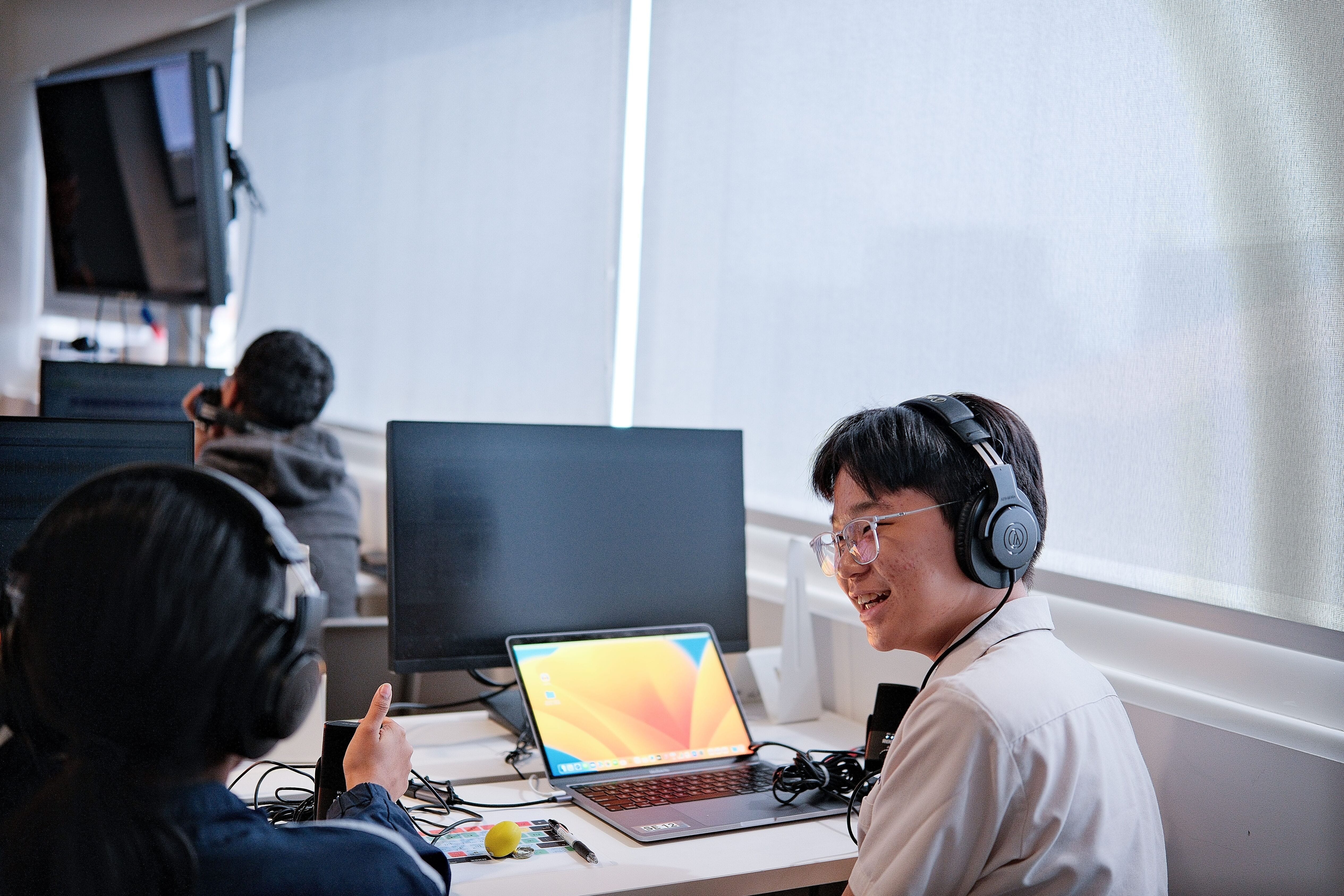 Two people wearing headphones talk at a desk with a laptop open, cables scattered, and monitors nearby.
