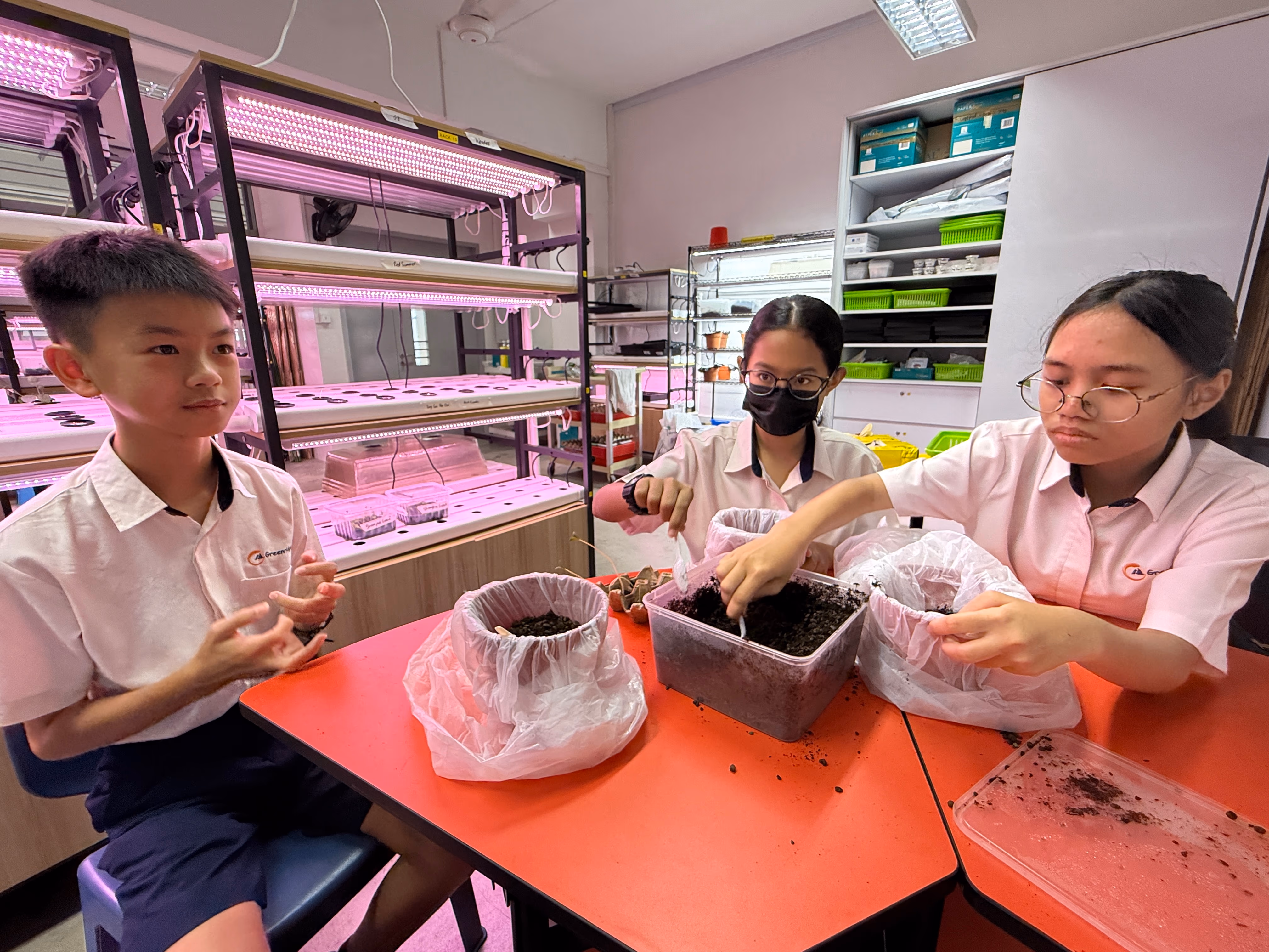 Three students in white uniforms work together on a cardboard model project at a table in a classroom.