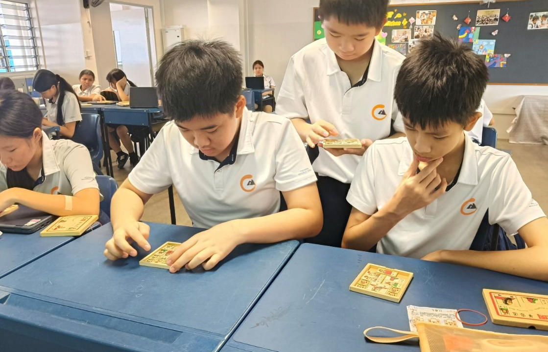 Three students in white uniforms examine small wooden puzzle tiles at blue desks in a classroom.