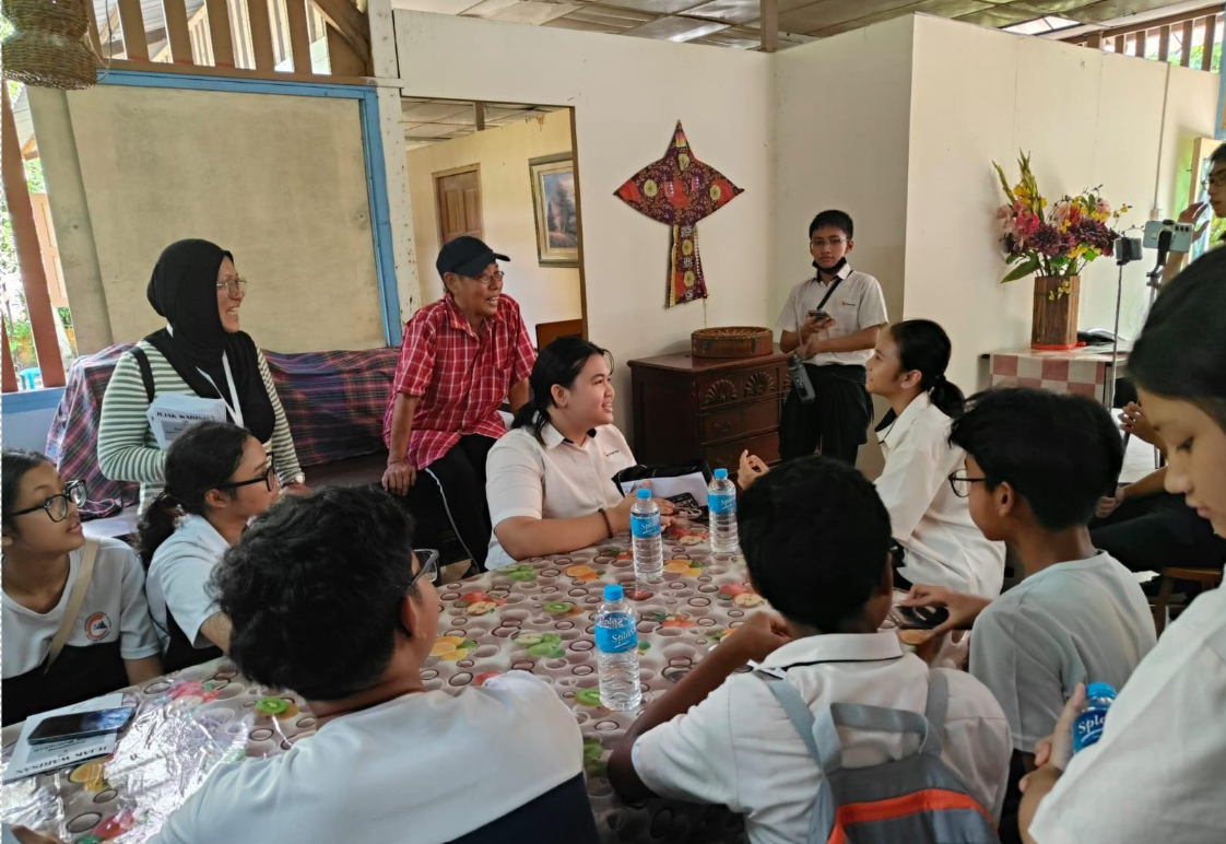 A group of students in white uniforms sit around a table with water bottles, talking with two adults indoors.