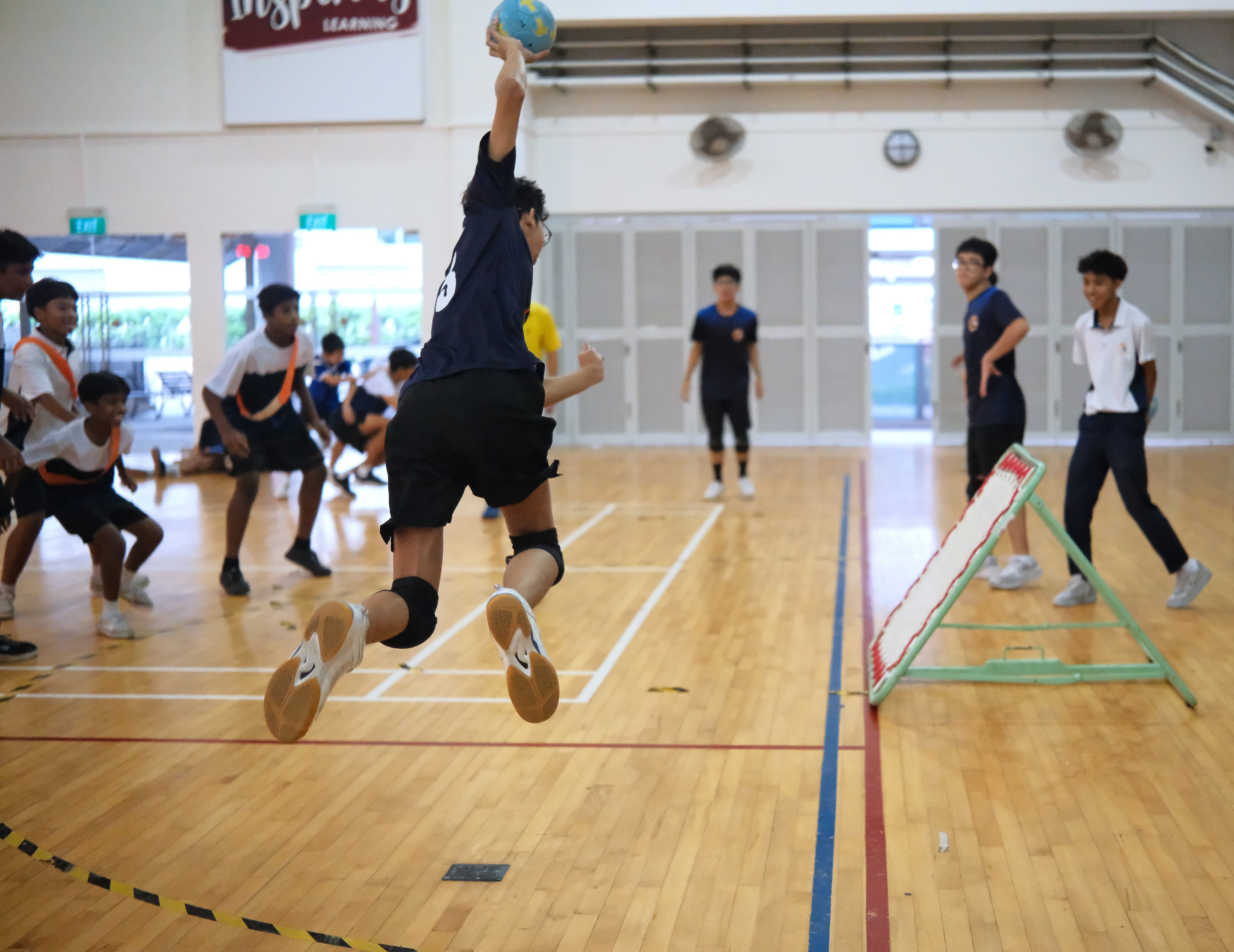 Student leaps in a gym holding a blue ball overhead as others watch near a small rebound net