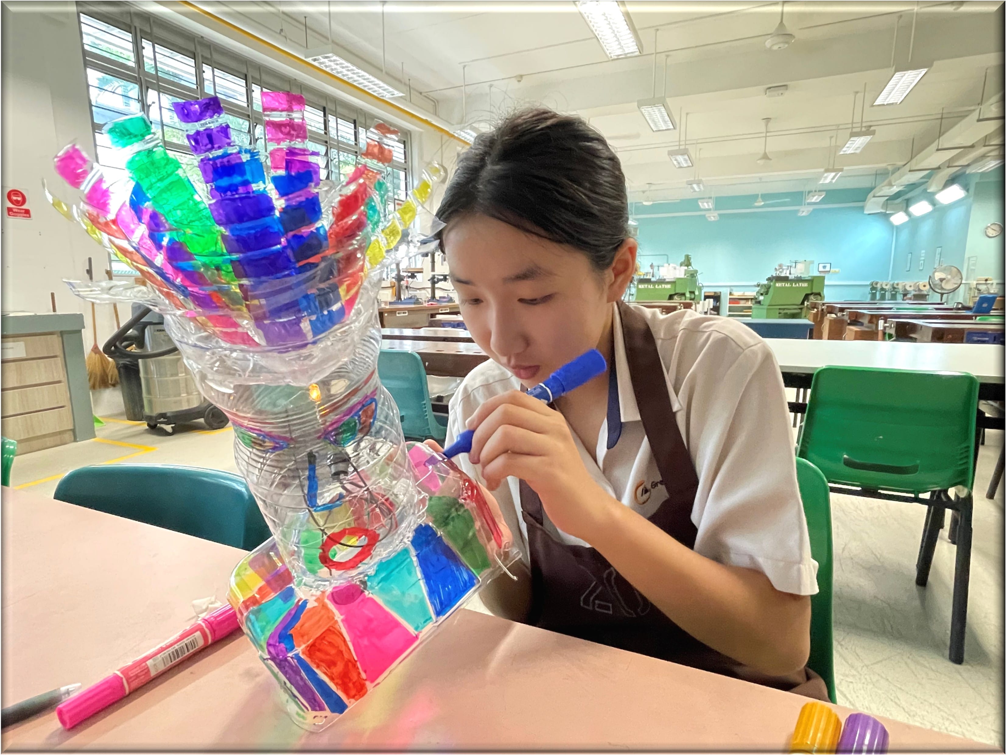 A young girl in a school apron colors a colorful plastic bottle sculpture with a blue marker in a workshop.