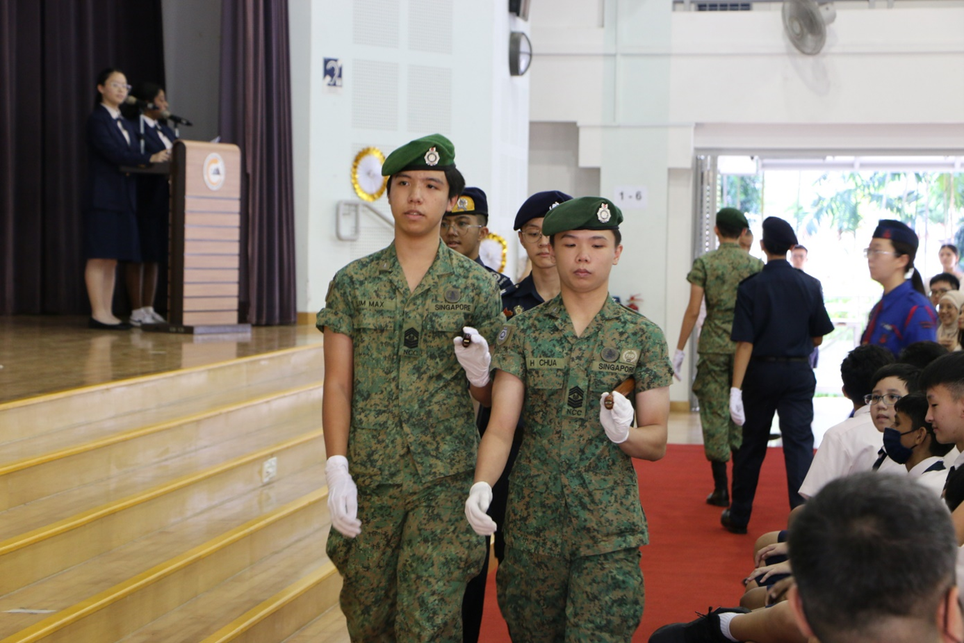 Two Singapore NCC cadets in camouflage uniforms and green berets march in formation during an indoor ceremony.