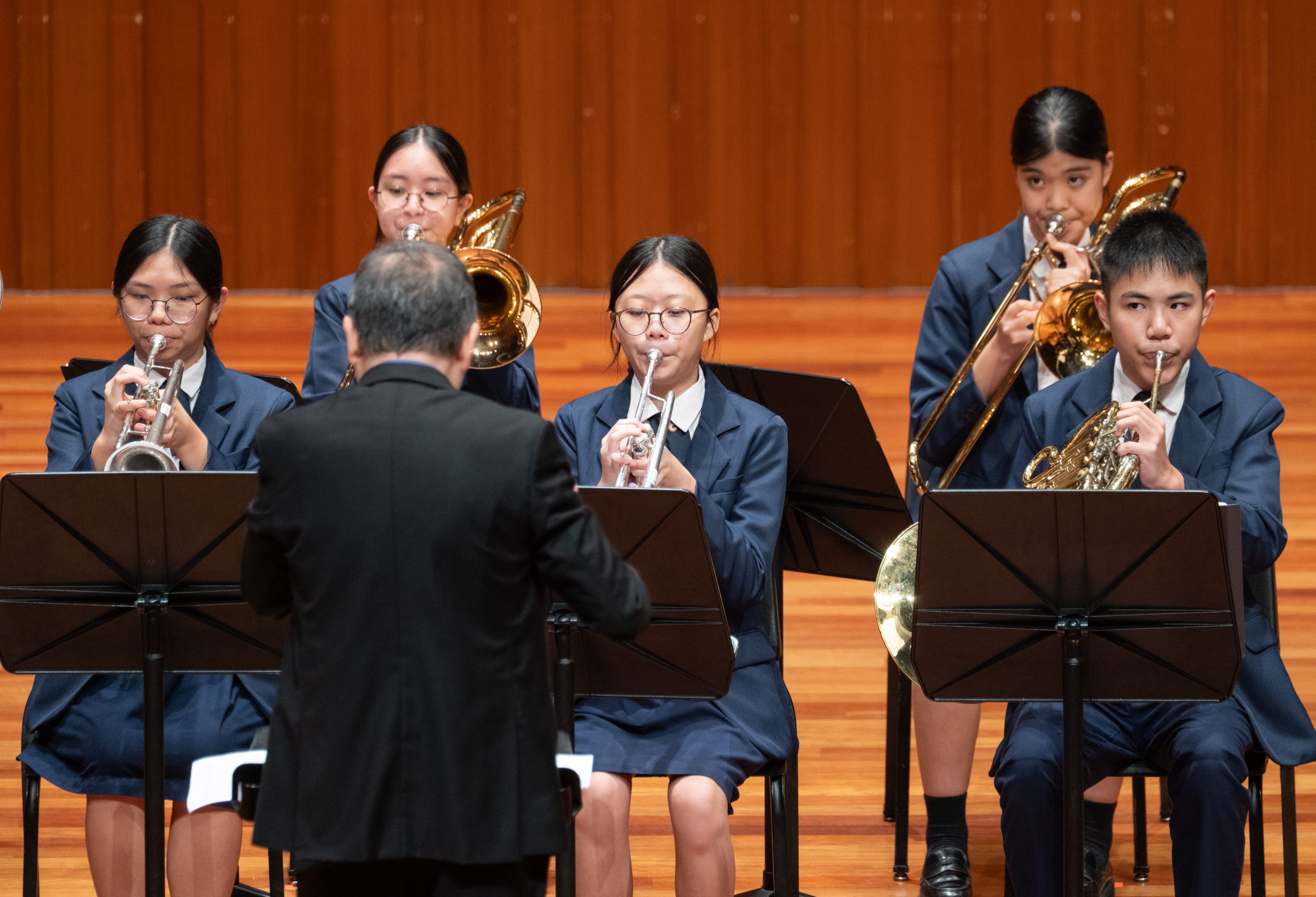 Five students in blue uniforms play brass instruments on stage, led by a conductor in black facing away.