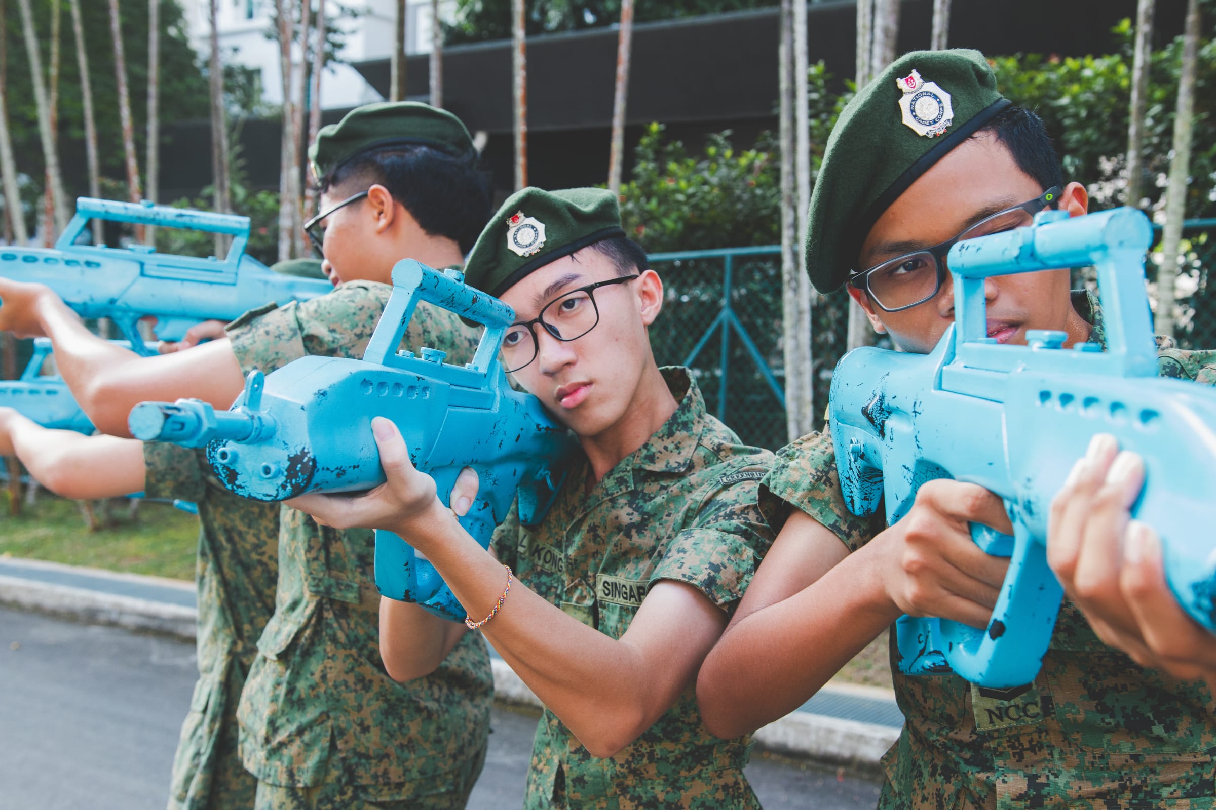 Three young cadets in camouflage uniforms and green berets aim blue training rifles outdoors.