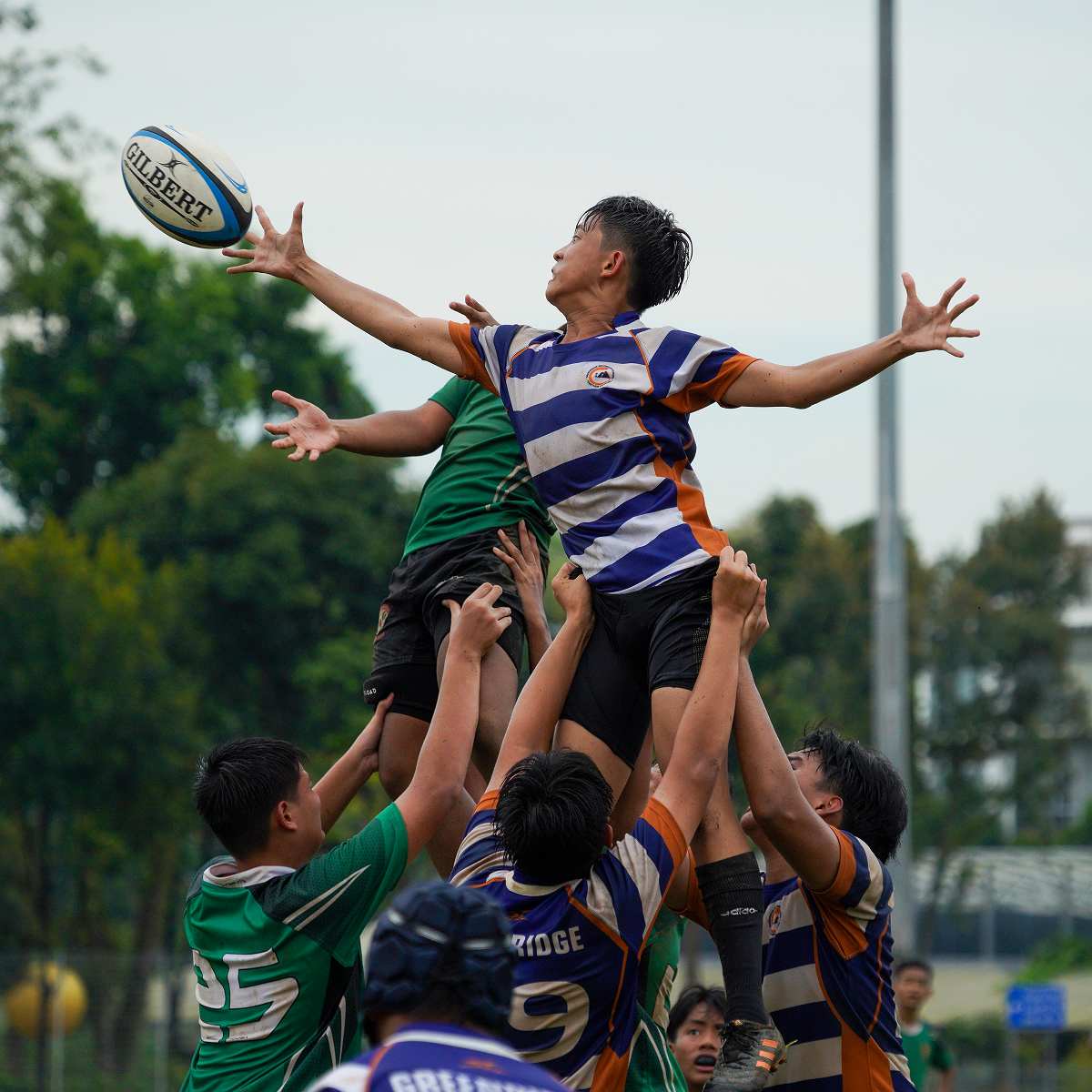 A rugby player in blue and white stripes is lifted by teammates to catch a Gilbert ball during a lineout.