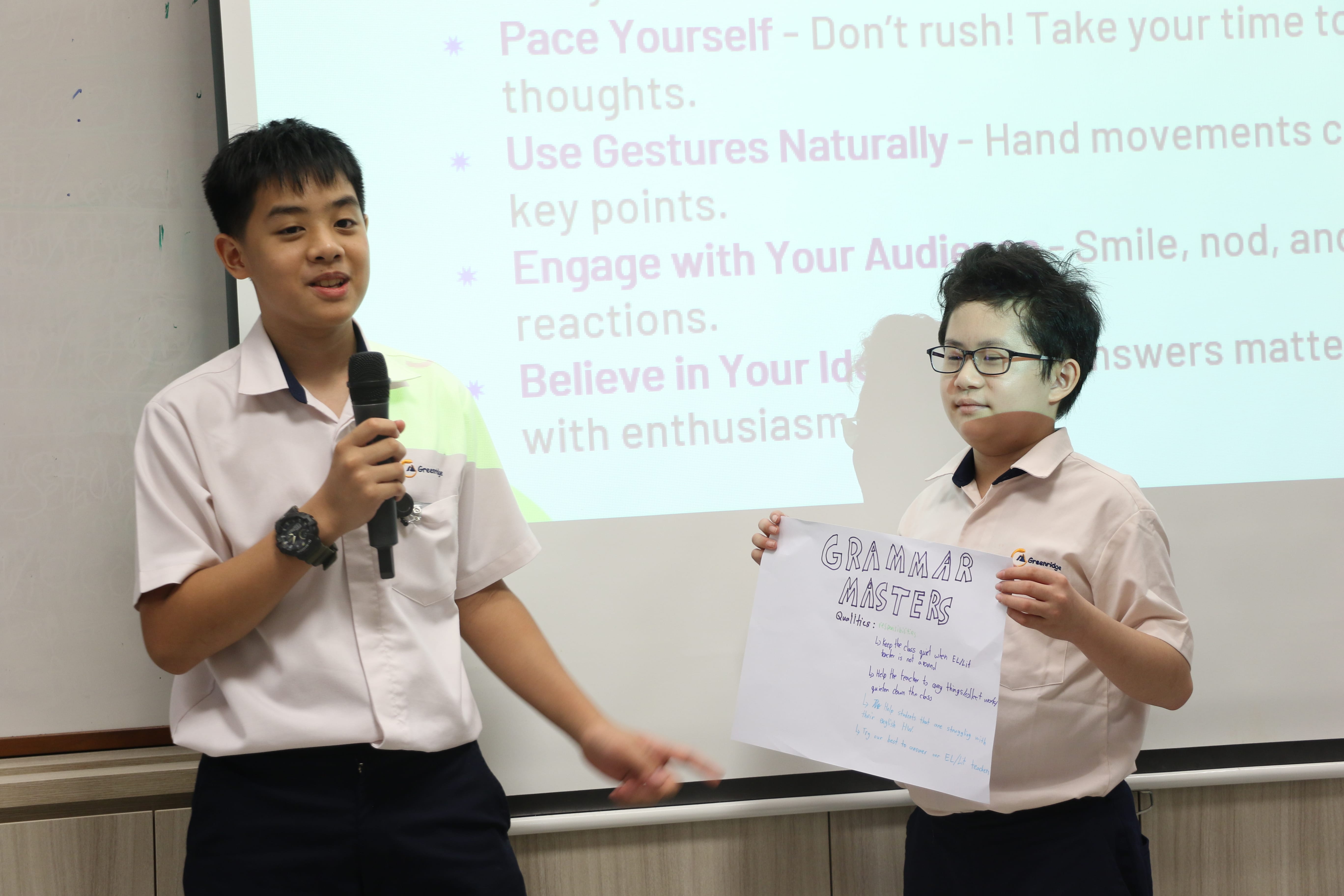 Student speaks into a microphone while another holds a Grammar Masters poster in front of a projector screen.