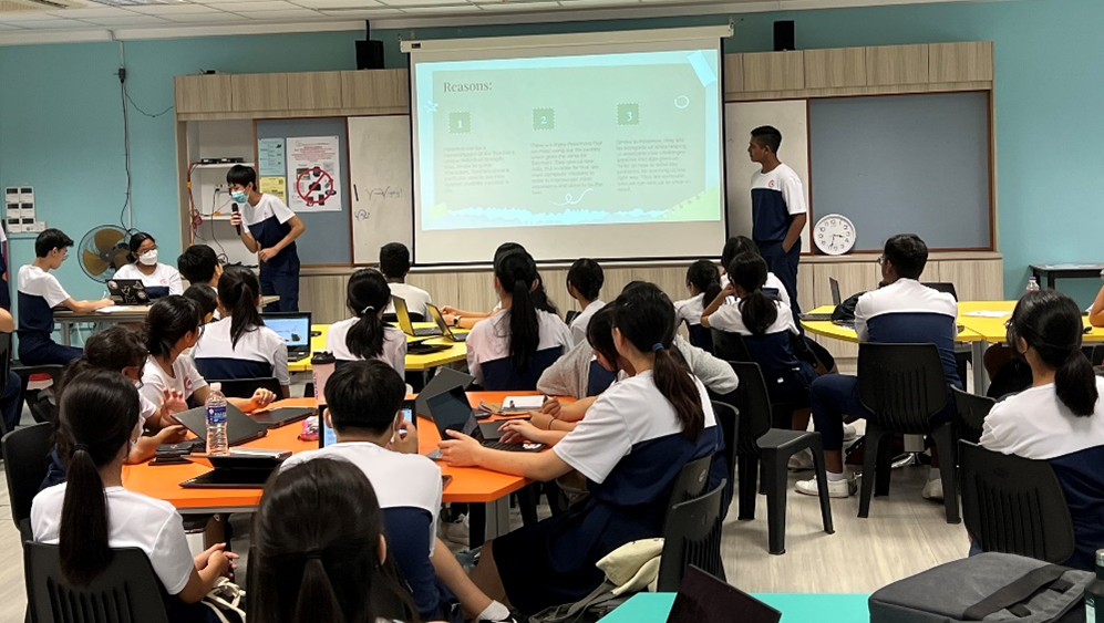 Students in uniforms sit at orange tables with laptops watching two classmates present a slideshow in a classroom.