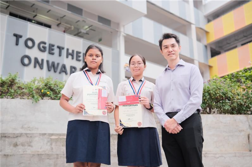 Two female students wearing medals hold certificates alongside a man, standing before a "Together Onwards" school sign.