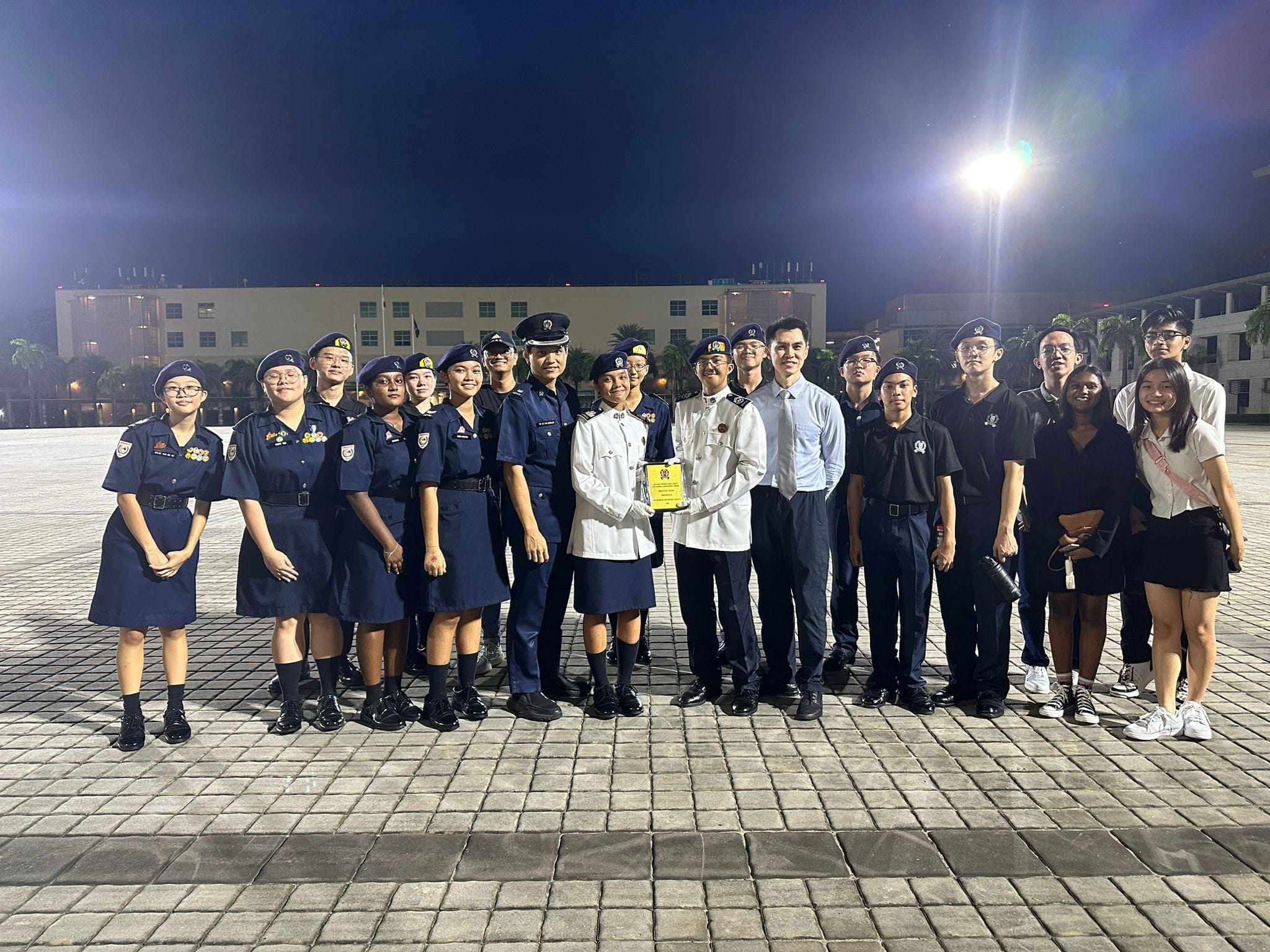 A group of young people in navy uniforms and white dress uniforms pose outdoors at night, two holding a yellow award plaque.