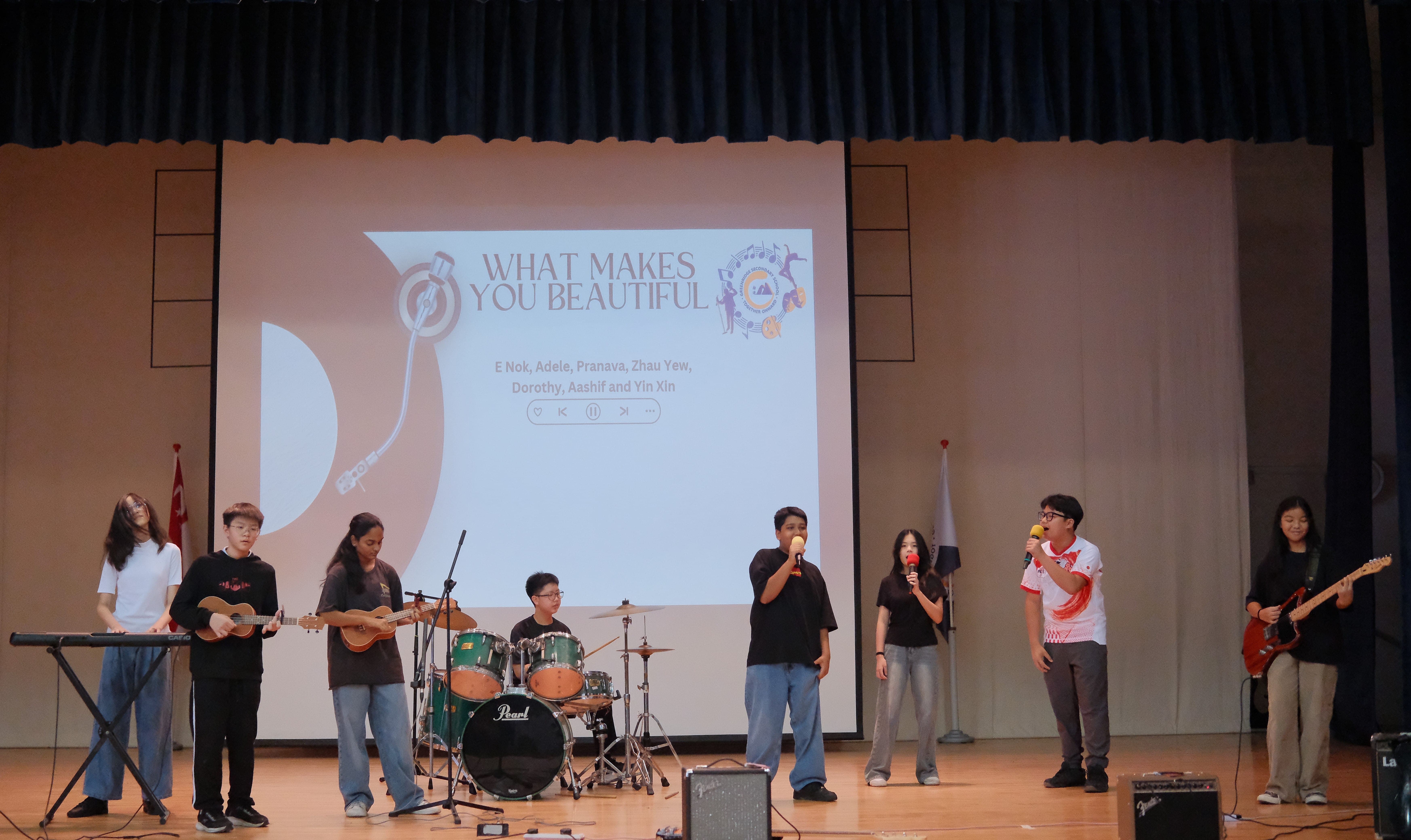 Student band on stage with drums, guitars, keyboard and singers; screen behind reads WHAT MAKES YOU BEAUTIFUL