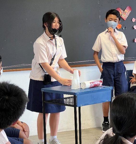 Two uniformed students present at a small table with cups in front of seated classmates near a chalkboard.