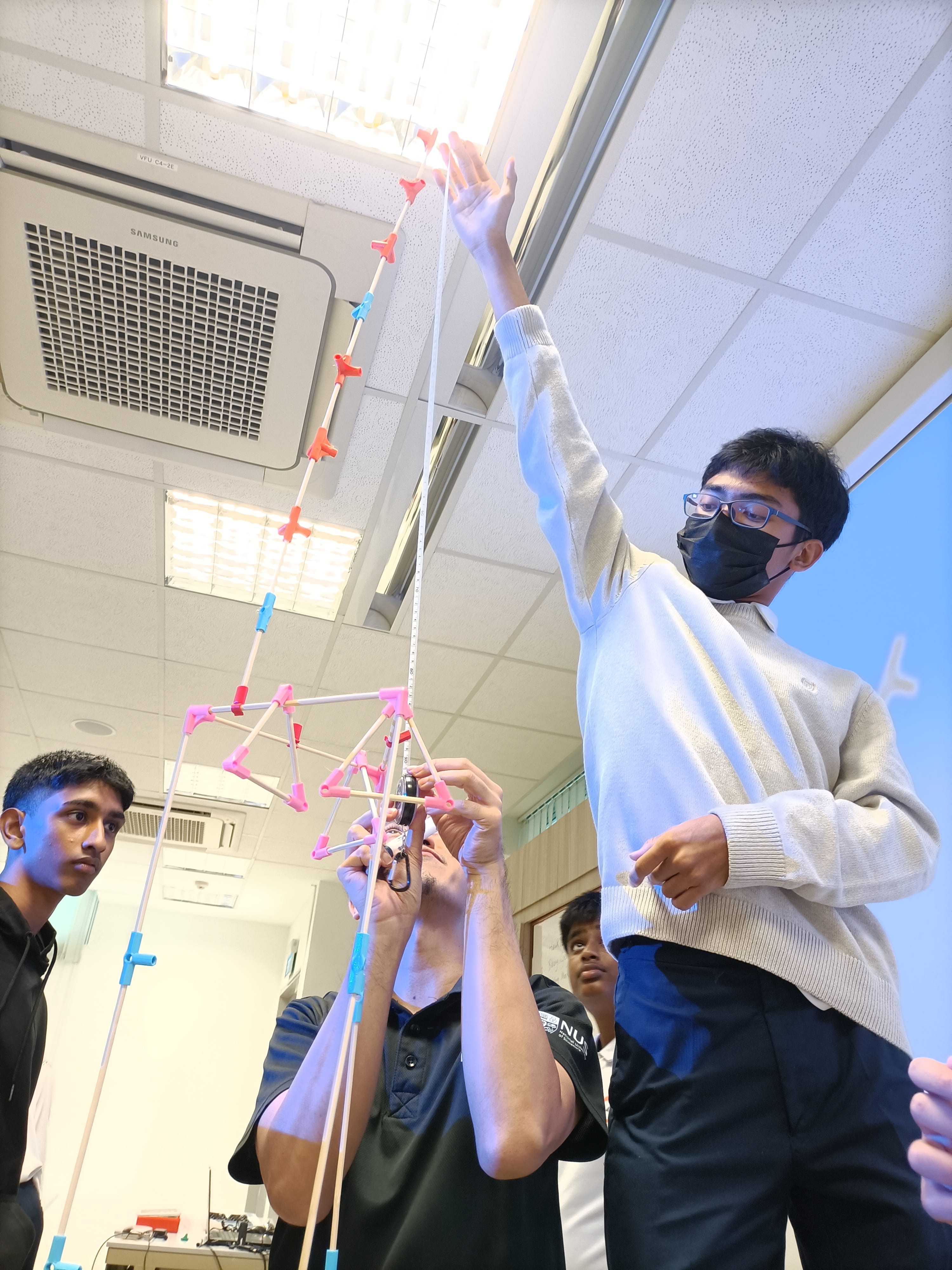 Students in a classroom hold up a tall colorful stick-and-connector structure while one measures its height.