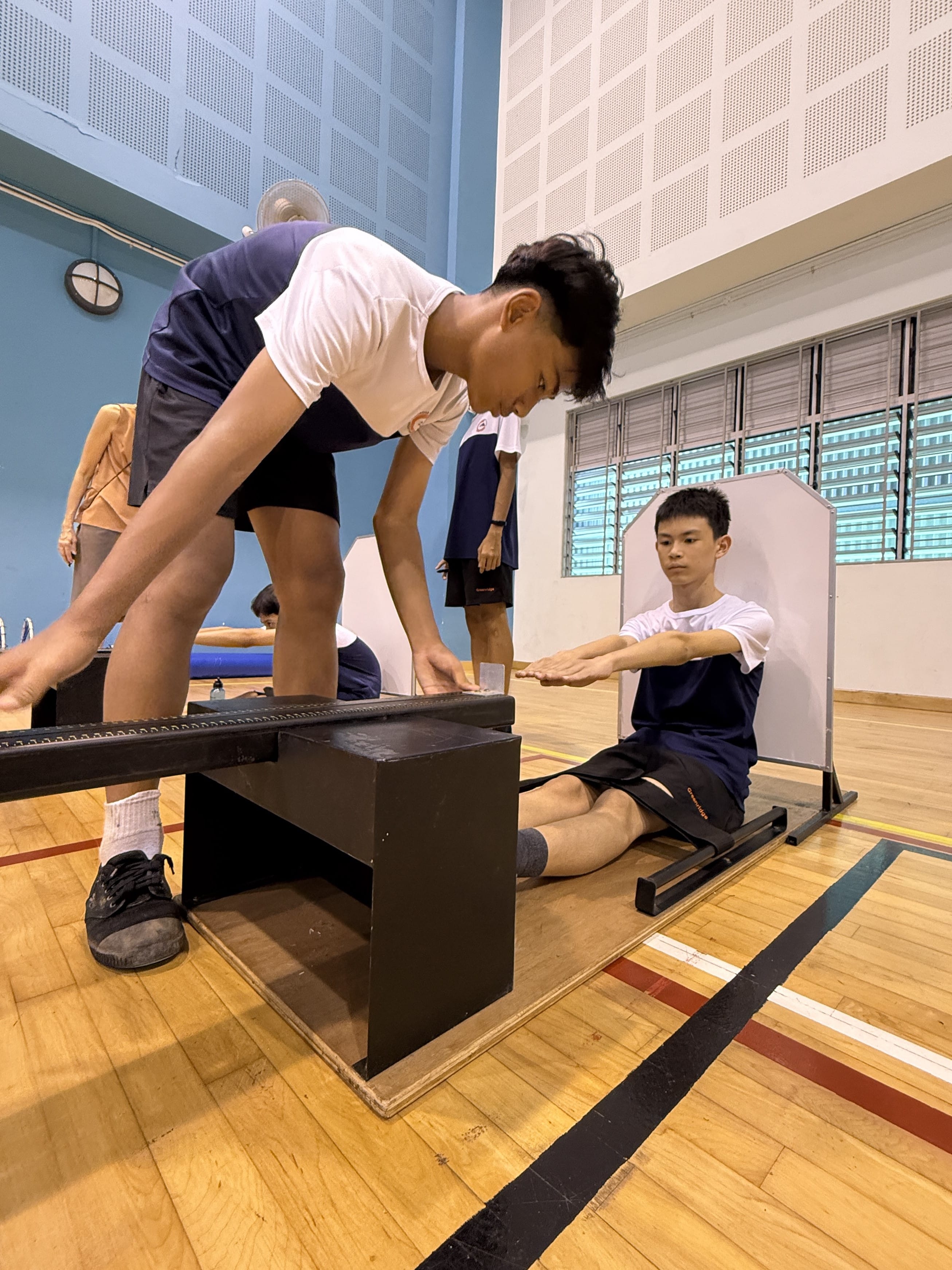 A teen sits on a sit-and-reach box stretching forward while another teen reads the measurement in a gym.