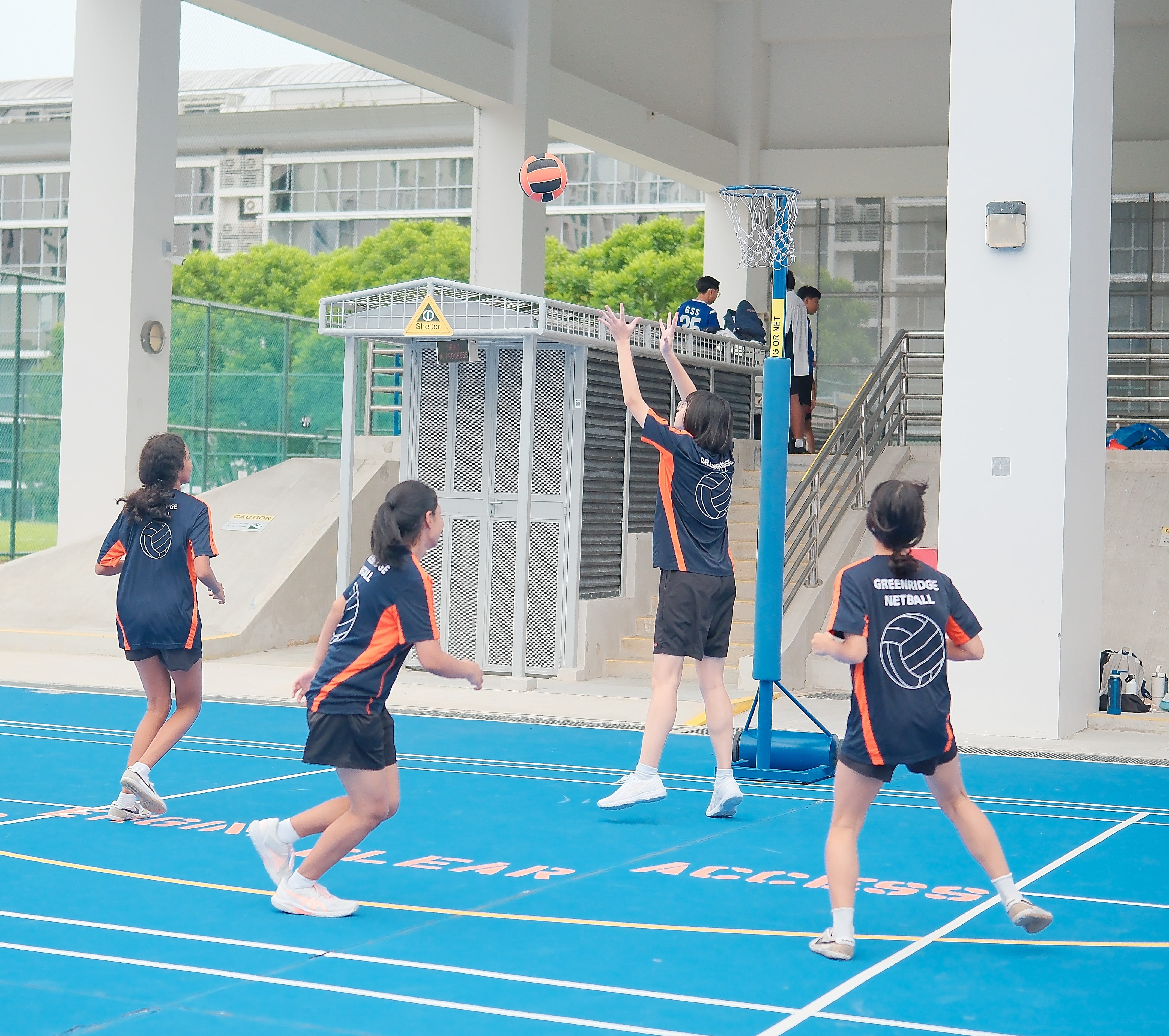 Four Greenridge Netball girls in navy and orange uniforms play netball on a blue outdoor court.