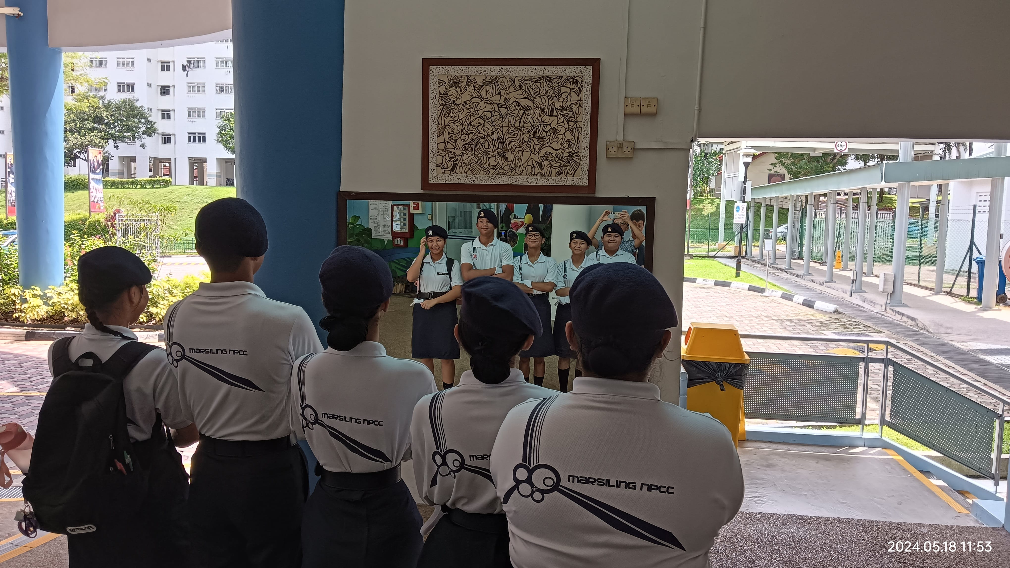 Students in Marsiling NPCC uniforms and berets gather outdoors, facing a group of peers posing in front of a mural.