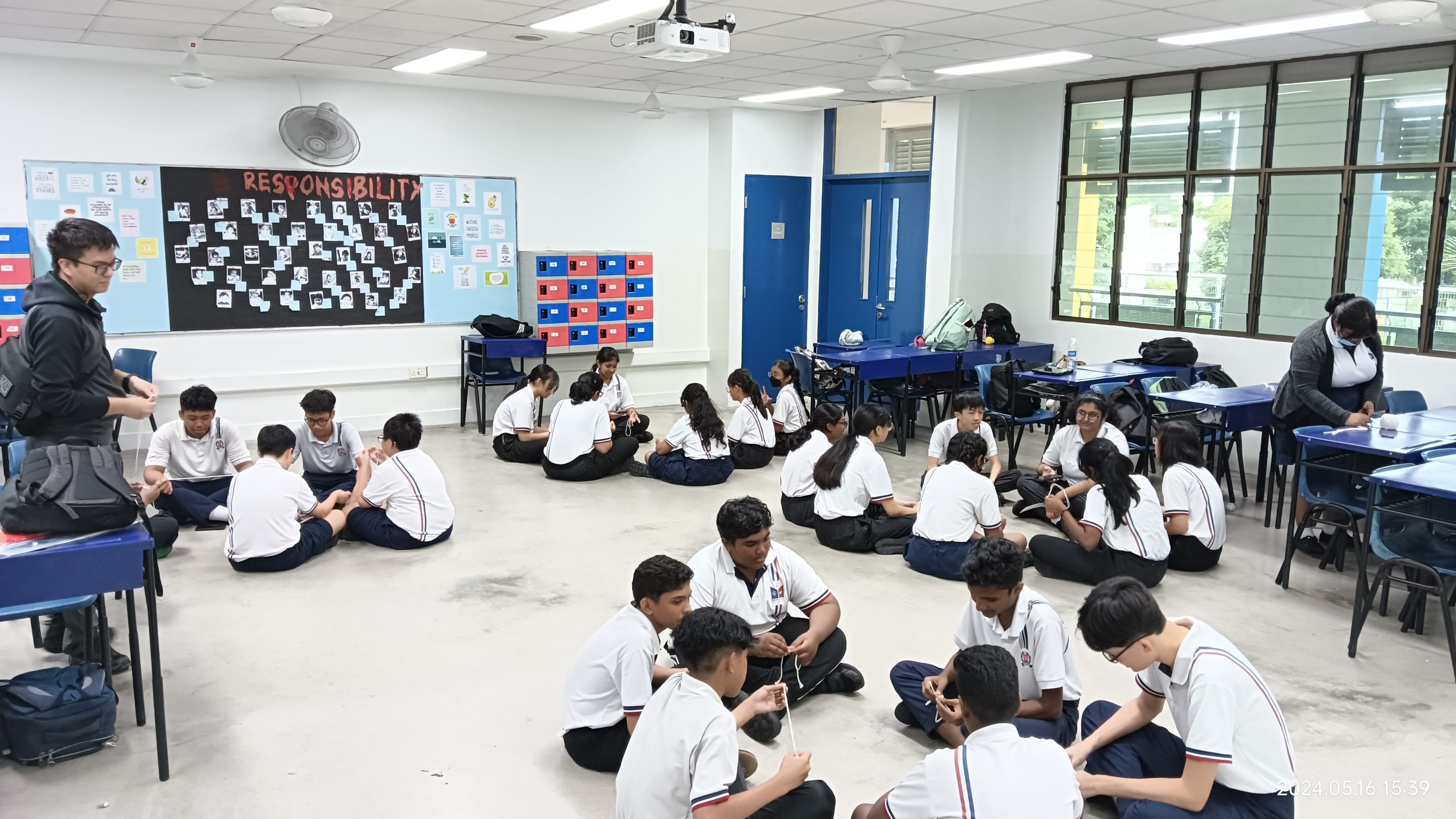 Students in white uniforms sit on a classroom floor in groups, with a Responsibility bulletin board on the wall.