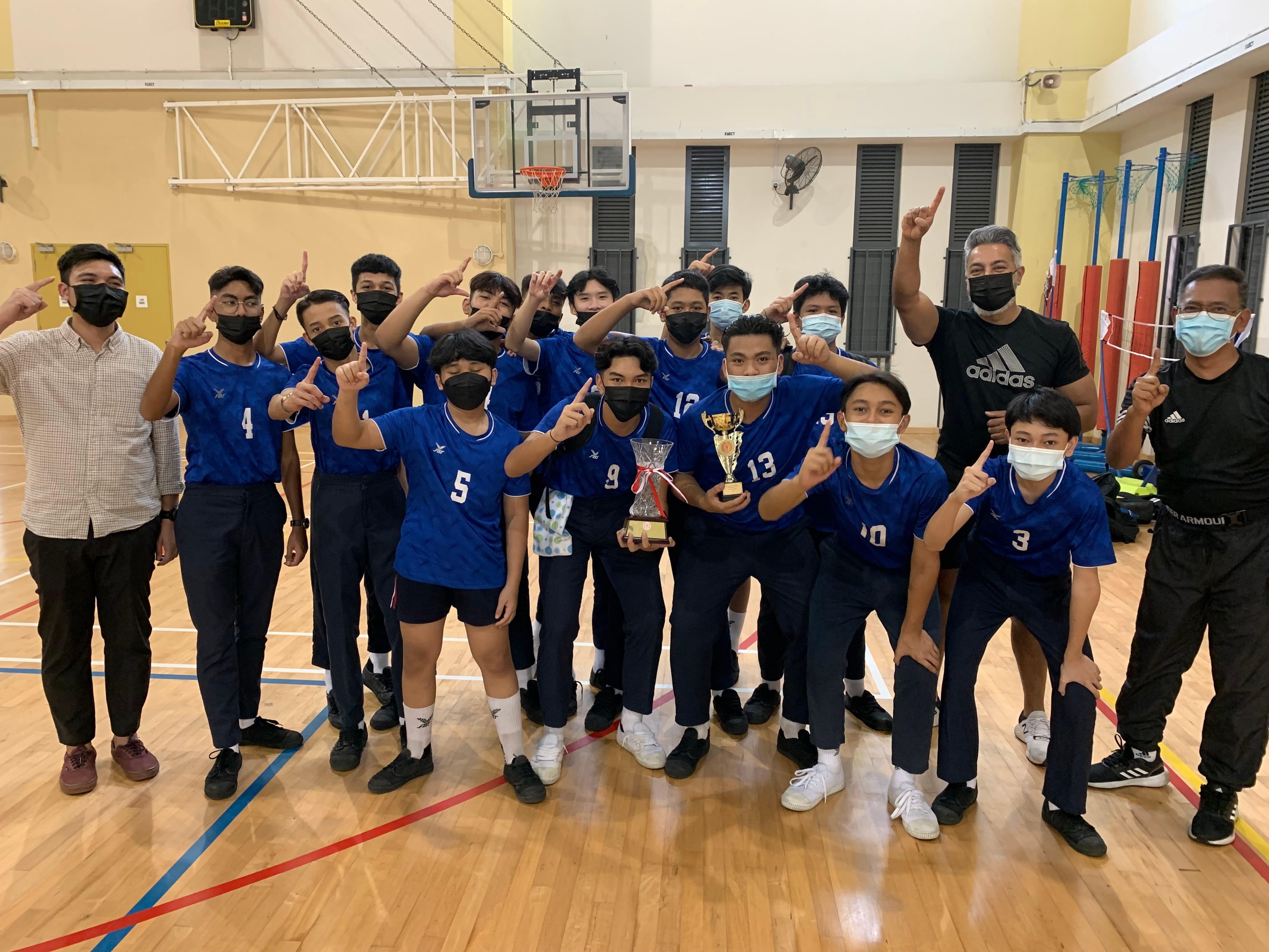 A masked youth sports team in blue jerseys poses with a trophy in a gymnasium, pointing fingers upward in celebration.