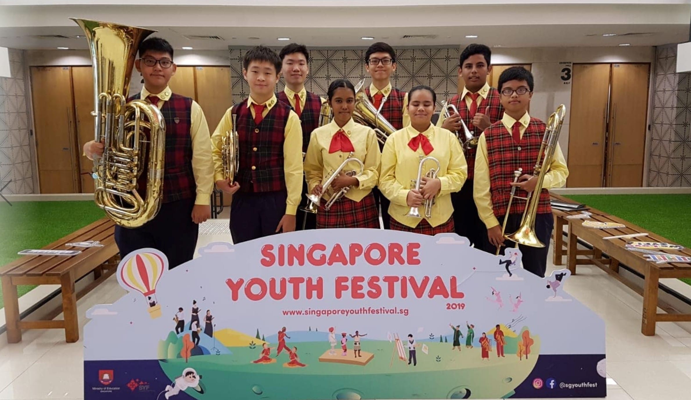 Seven students in matching uniforms hold brass instruments behind a Singapore Youth Festival 2019 sign.