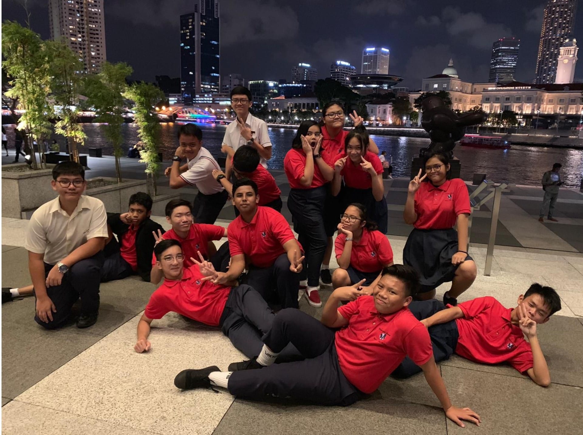 A group of teenagers in red uniforms pose playfully along a lit city waterfront at night.