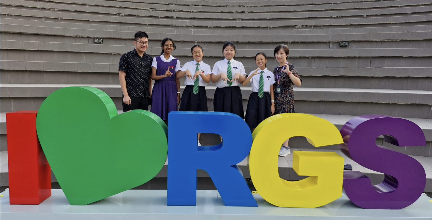 Six people pose behind large colorful letters spelling "I heart RGS" in an outdoor amphitheater setting.