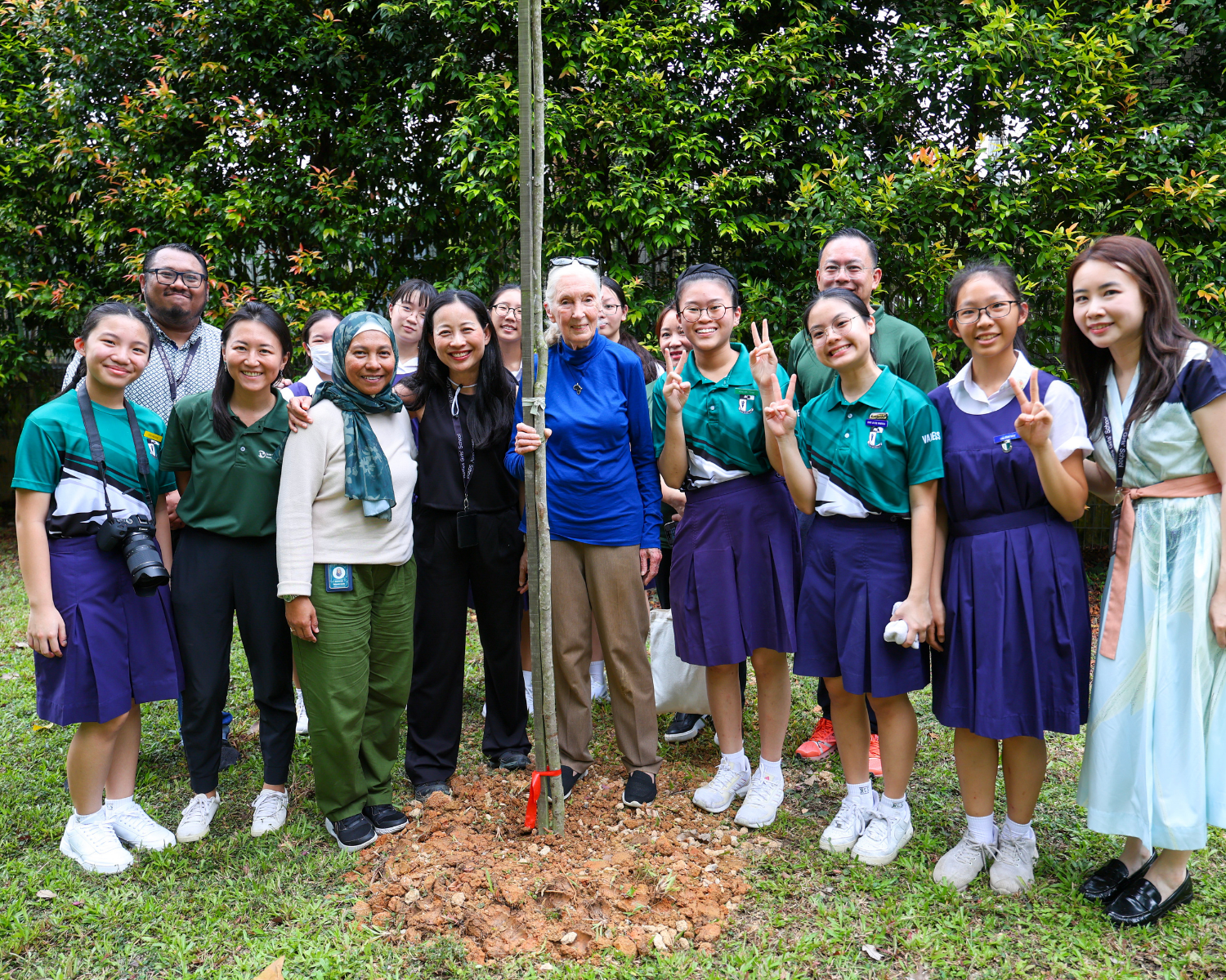 Group posing outdoors by a newly planted tree; several students in uniforms flash peace signs.