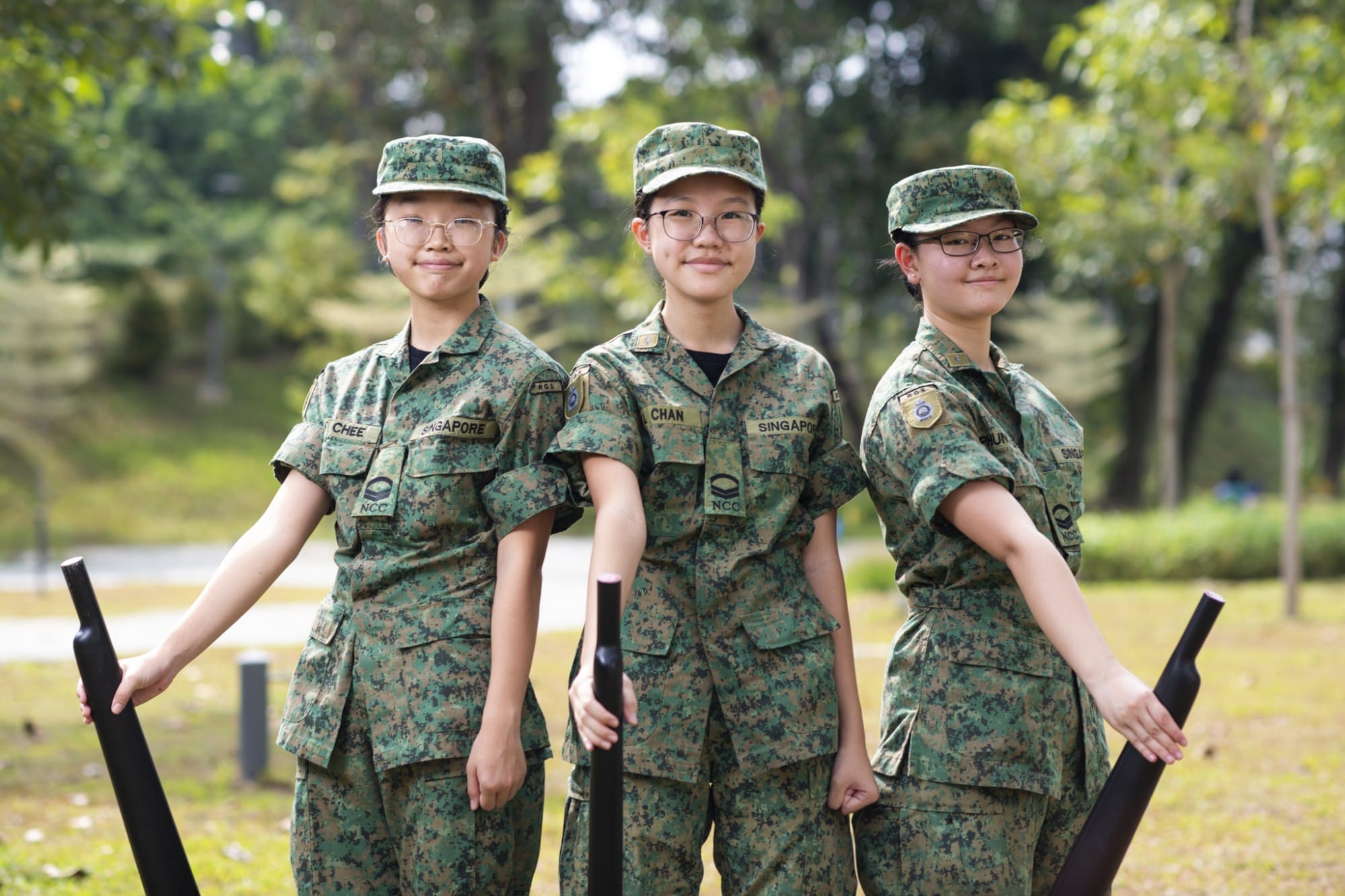 Three young women in Singapore NCC camouflage uniforms and caps smile outdoors, each holding a black mock rifle.