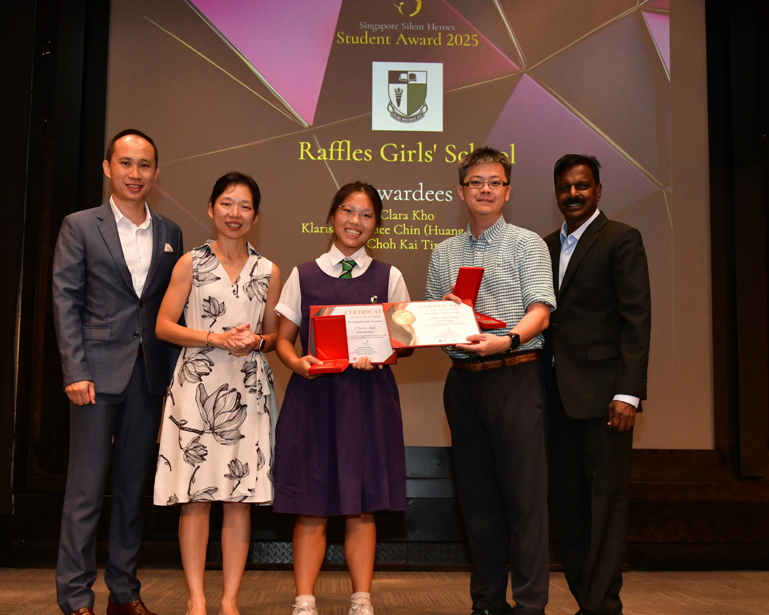 Five people pose on stage at the Singapore Silent Heroes Student Award 2025 for Raffles Girls' School, holding certificates.