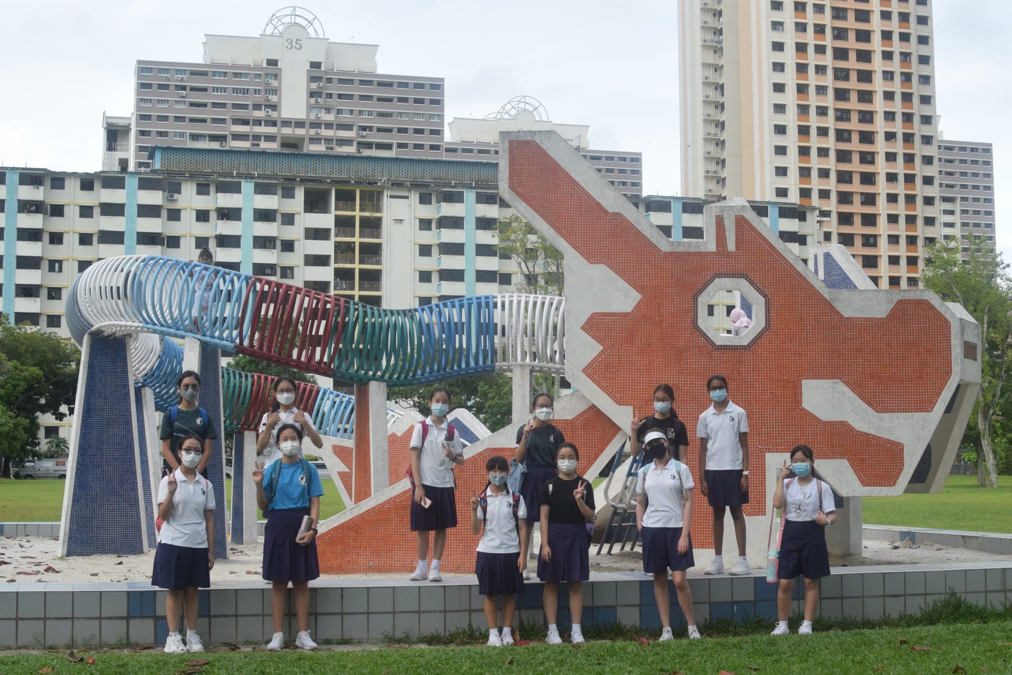 Students in uniforms and masks pose in front of a large dragon-shaped mosaic playground with tall apartment blocks behind.