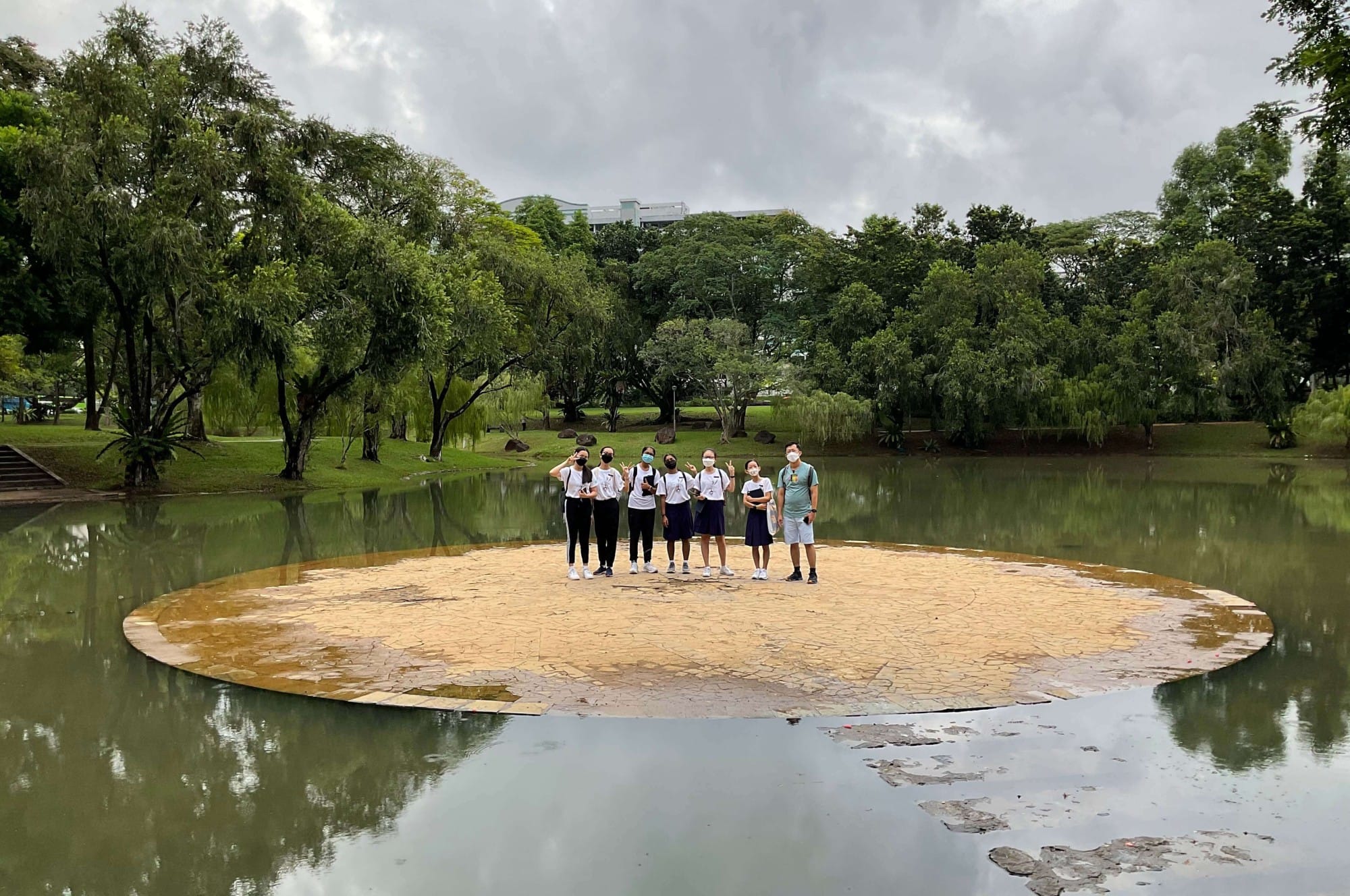 Six masked people stand on a circular stone platform in a pond, surrounded by lush green trees under a cloudy sky.