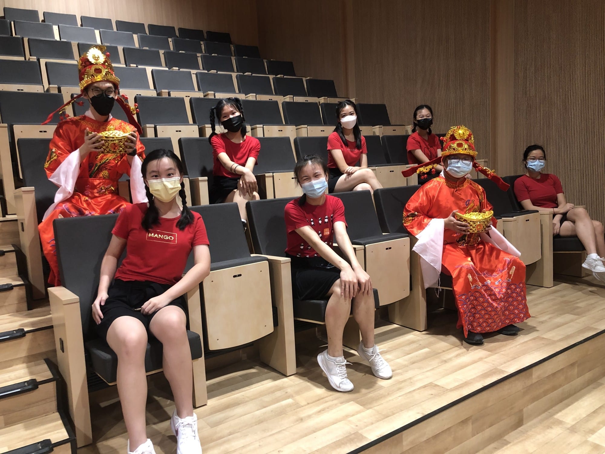 Seven masked people sit in an auditorium; two wear red Chinese traditional costumes with gold headdresses.