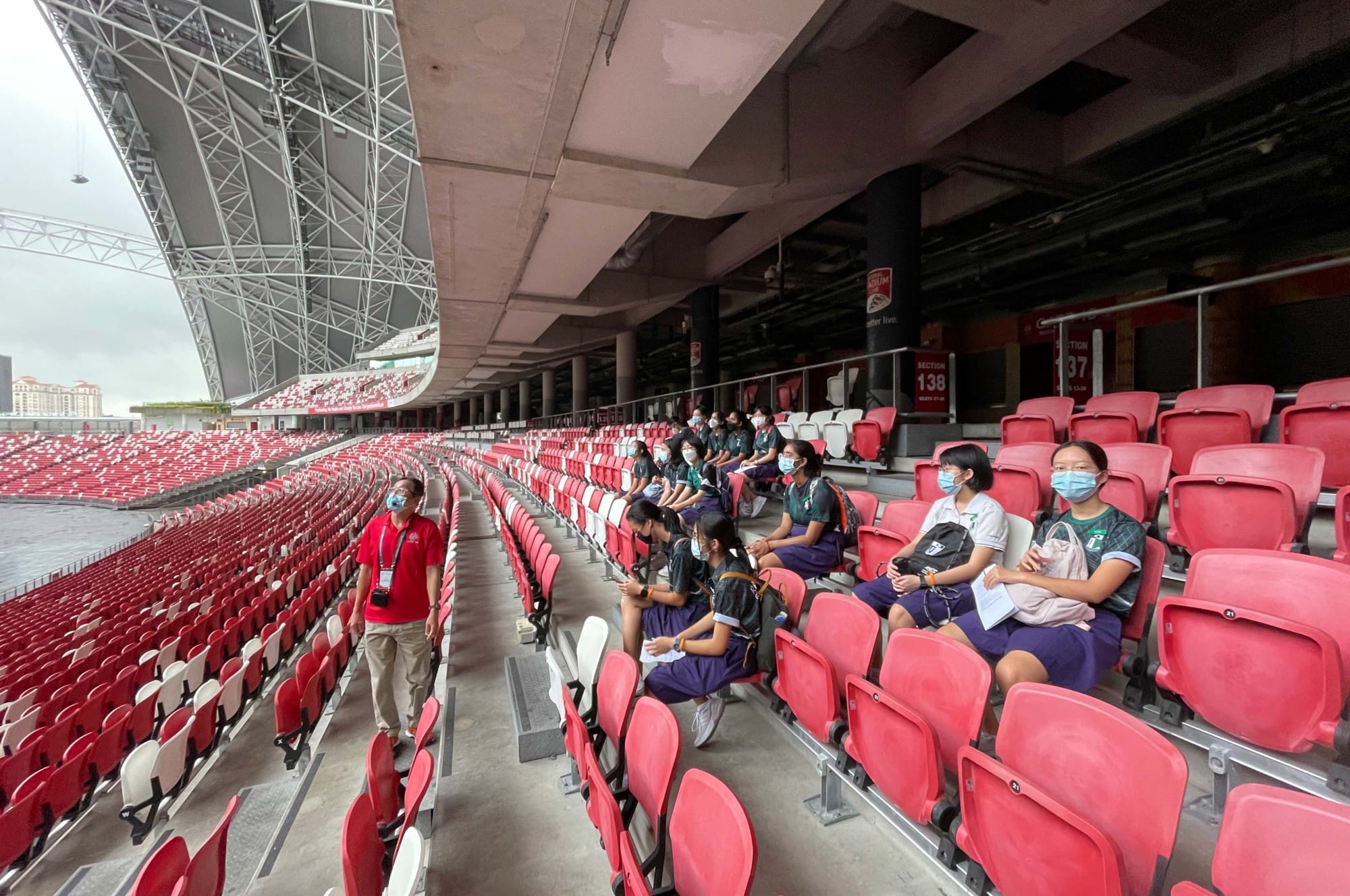 Students wearing masks sit in red stadium seats while a guide in red walks along the aisle during a tour.